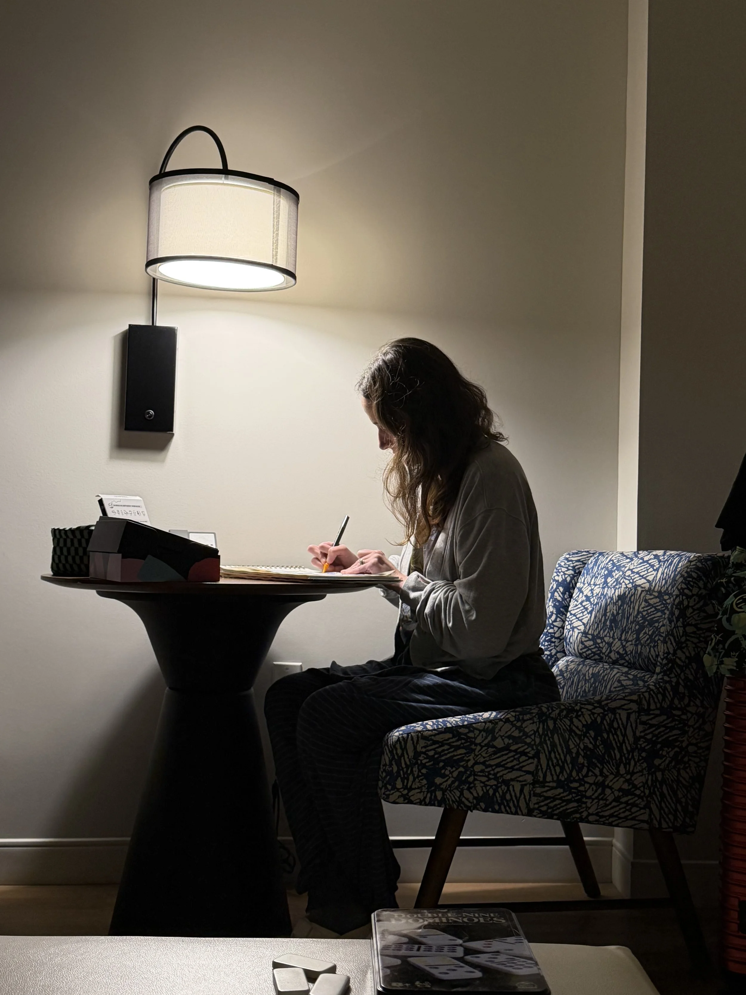 A woman with wavy brown hair sitting at a small round table, writing on a notepad. There is a black box with a checkered pattern, some papers, and a pen on the table. Behind her is a patterned armchair and a wall with a modern wall-mounted light fixture illuminating the scene.