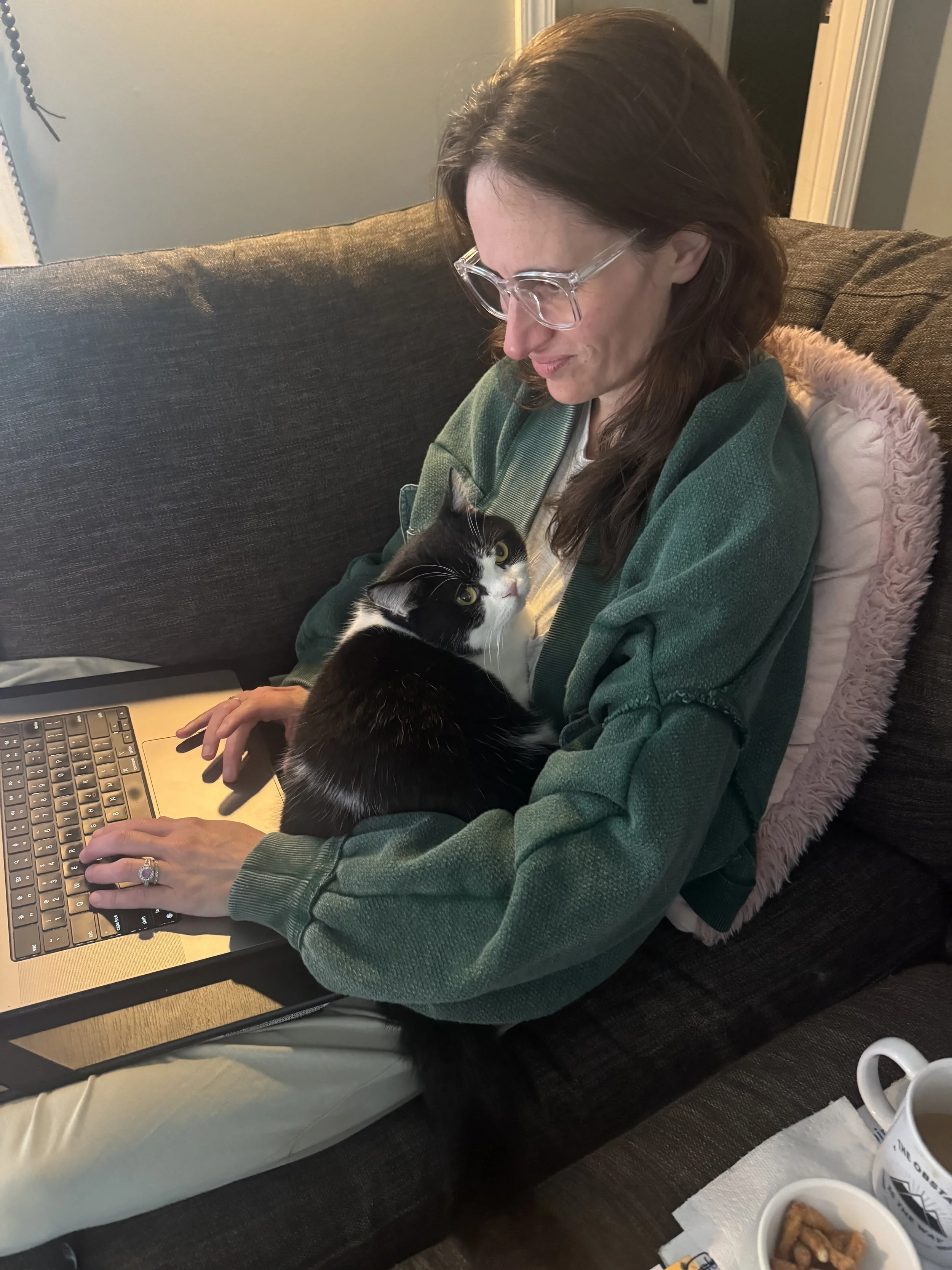 Meredith Rodkey with glasses and long brown hair sitting on a dark gray couch, working on a laptop, with a black and white cat sitting on her lap looking up at her in a cozy living room.