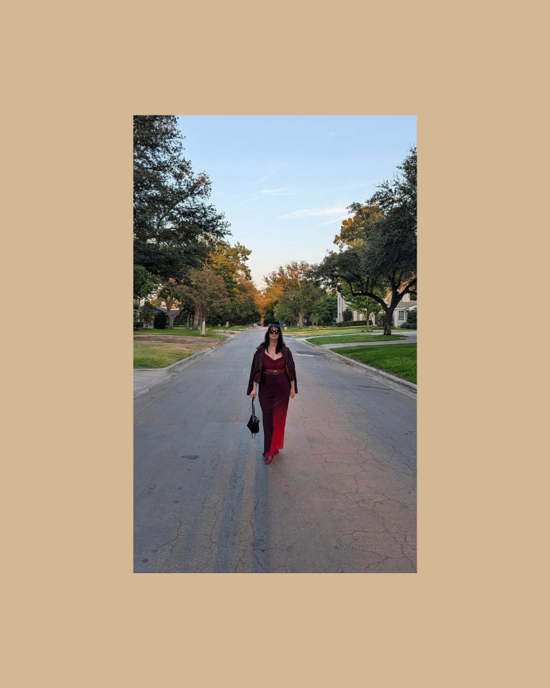 A woman walking down a quiet residential street during daytime, surrounded by trees and houses, wearing a red and black outfit and carrying a small handbag.