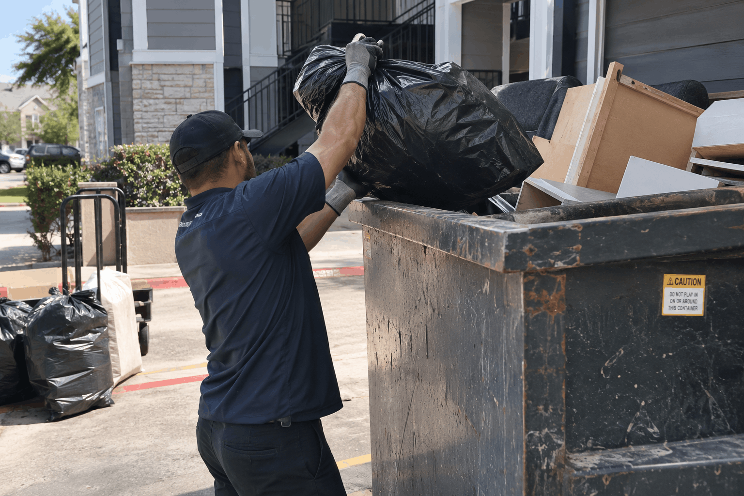A man wearing a dark blue uniform and a black cap is throwing a black trash bag into a large black dumpster.