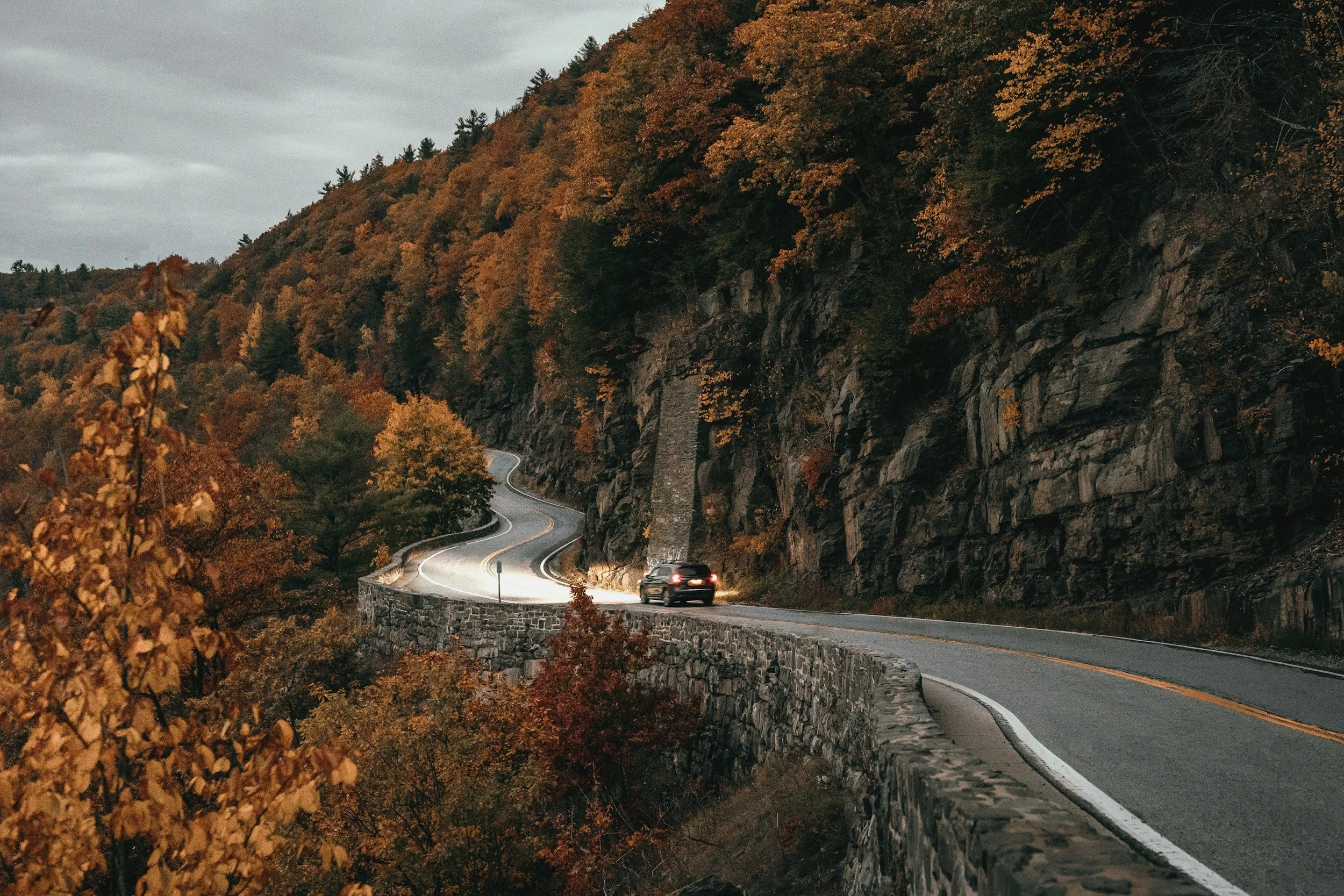 A winding mountain road with a car driving on it, surrounded by colorful autumn foliage and rocky cliffs under a cloudy sky.