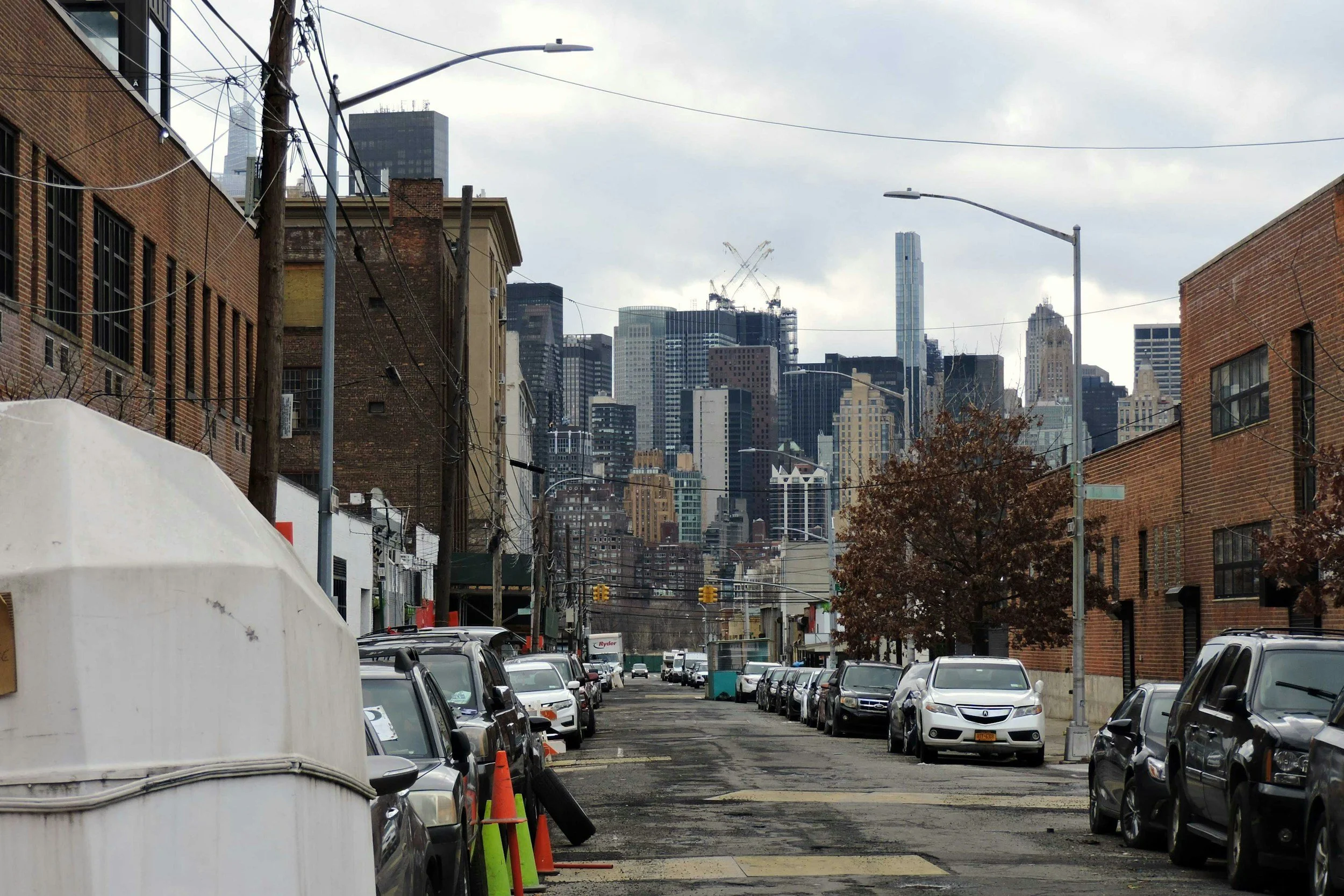 City street view with parked cars, utility poles, and high-rise buildings in the background on a cloudy day.