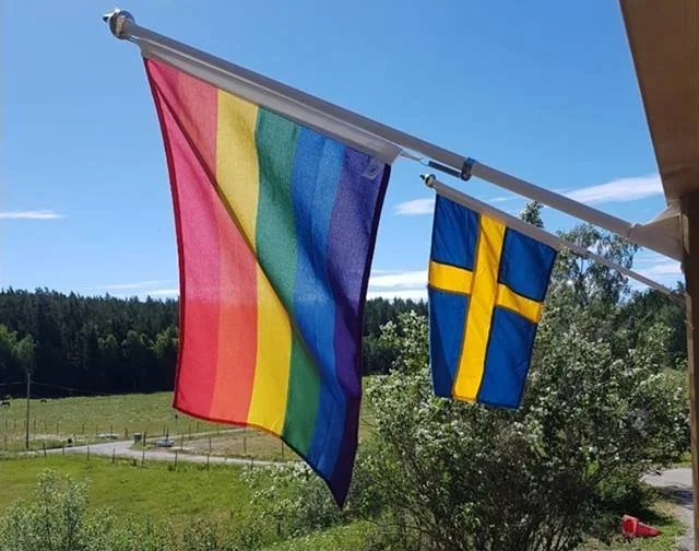 Två flaggor, en med regnbågsfärger och en med Sveriges flagga, hänger utomhus mot en blå himmel och gröna träd.