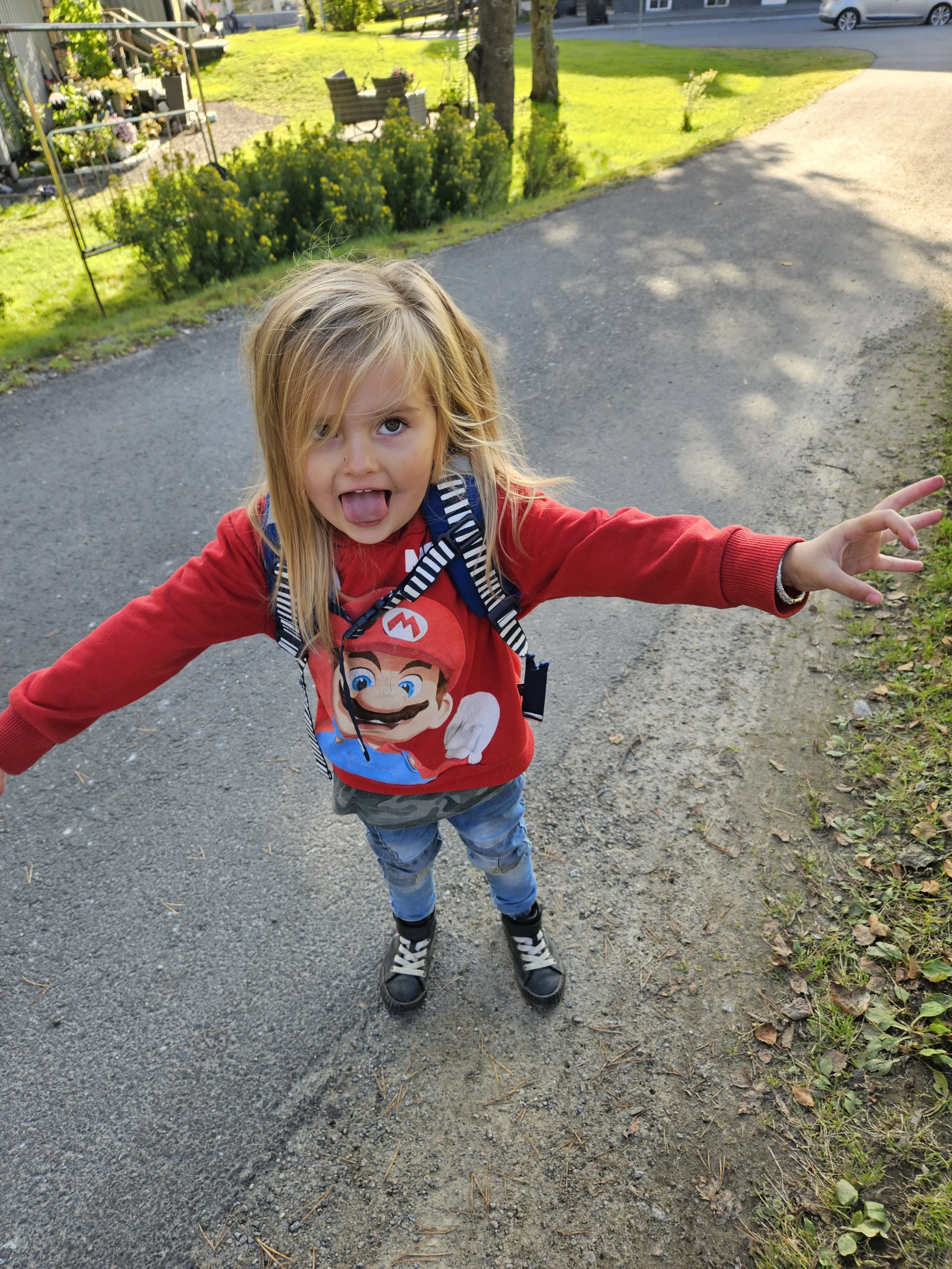 En ung flicka i röd tröja med Mario-figurtryck gör ett leende och sträcker ut armarna mot kameran, stående på en gångväg med gräs och träd i bakgrunden.
