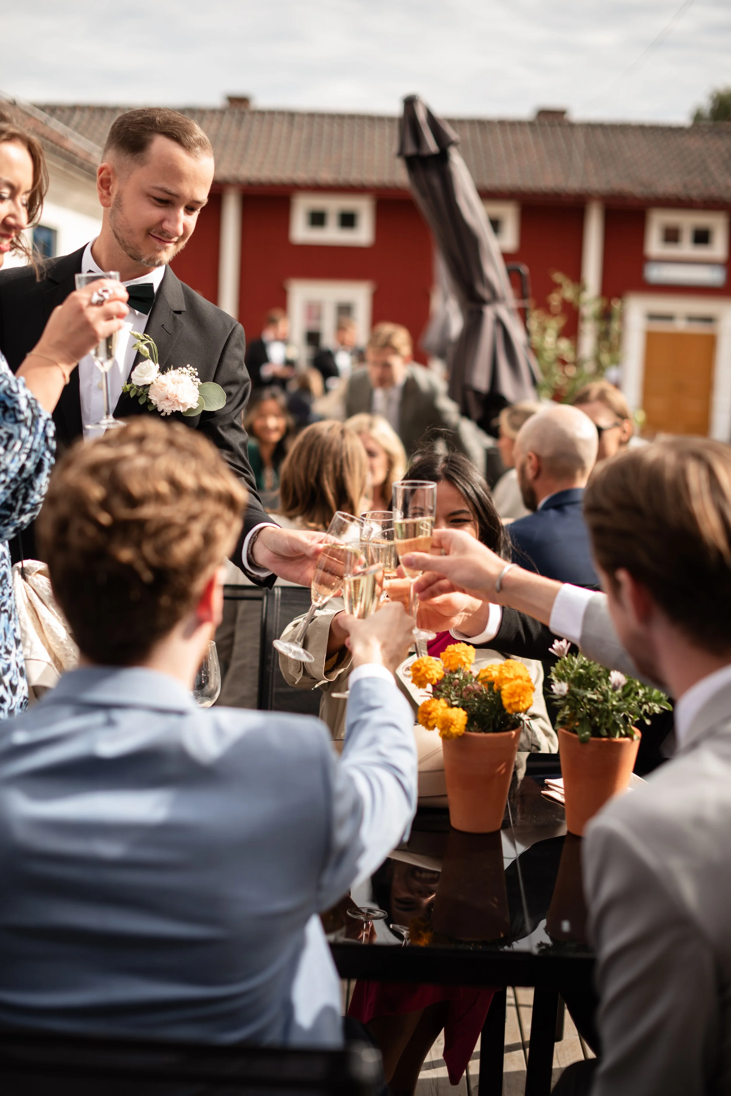 Före detta brudpar som utbyter skålar vid ett bord under ett bröllop utomhus, med många gäster och ett rött hus i bakgrunden.