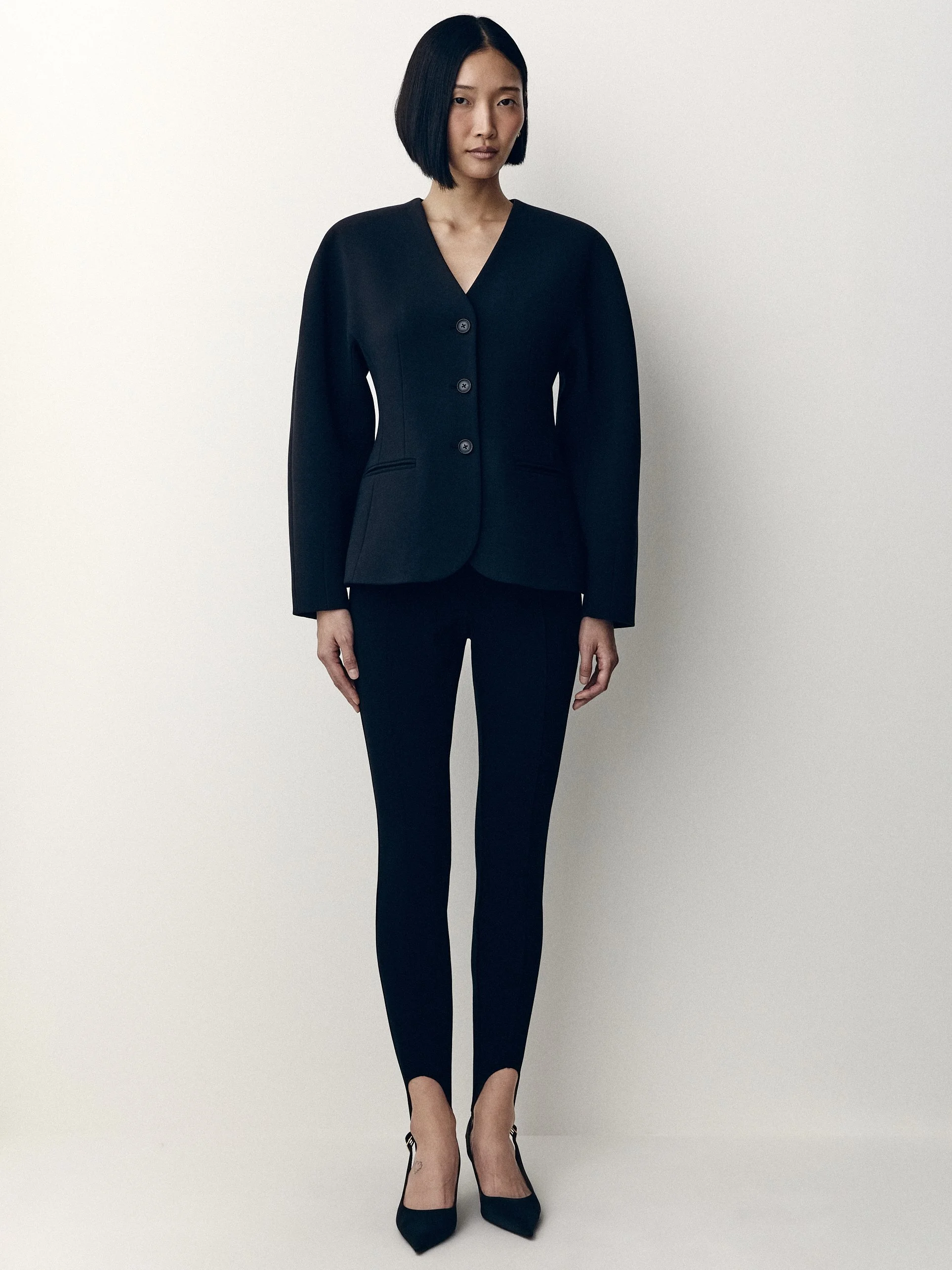 Woman facing the camera in a black cardigan, cropped black trousers and pointed heels.