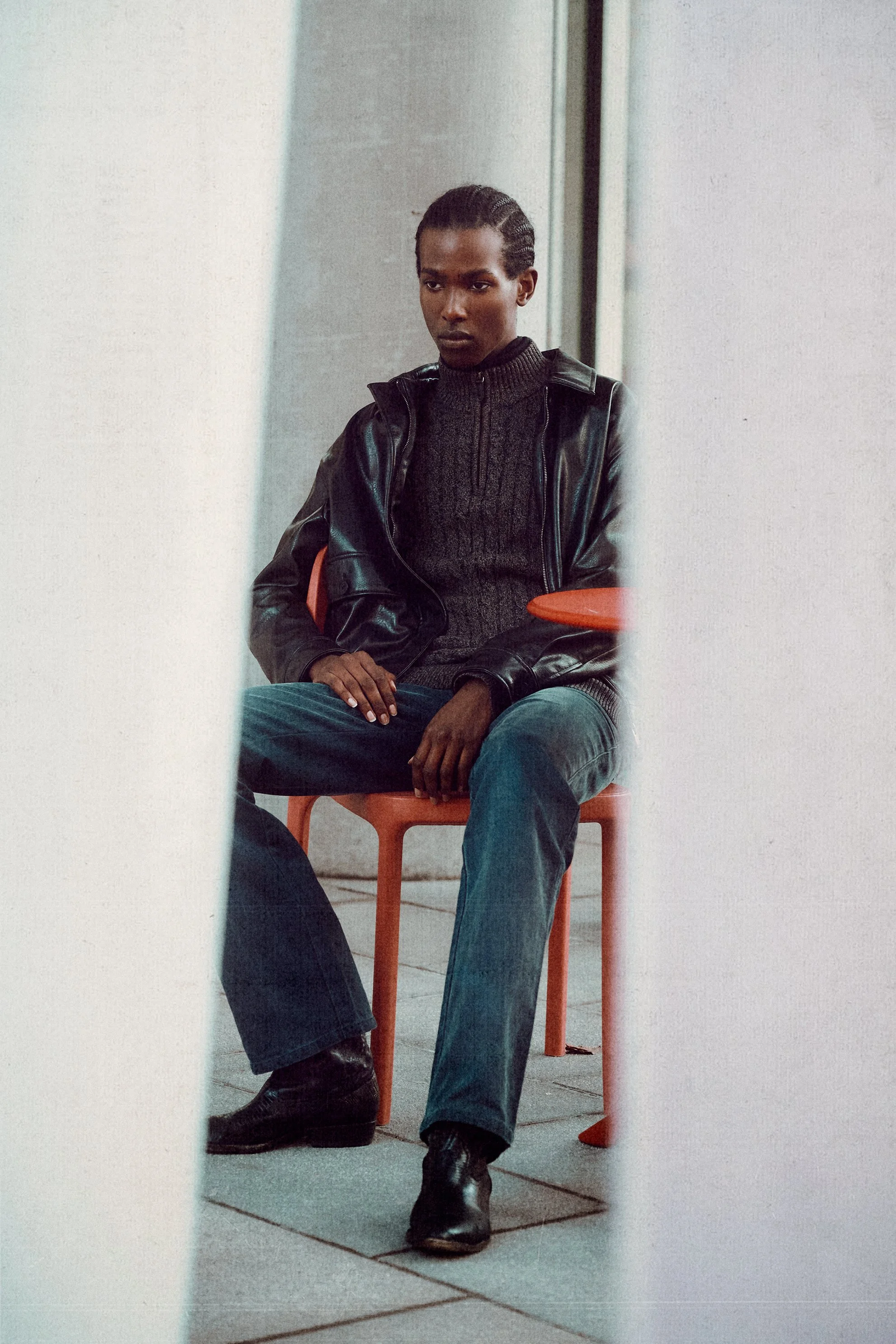 Man seated on a chair between pale columns wearing a leather jacket, dark sweater and jeans.