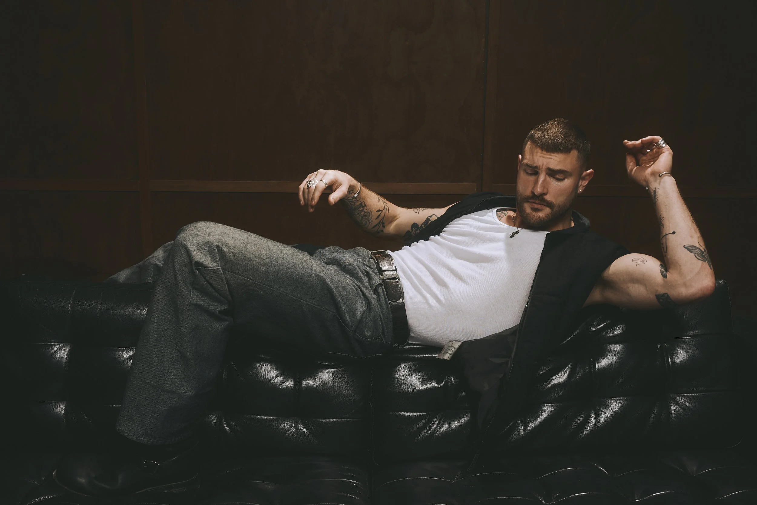 Tattooed man reclining across a leather sofa in a white tank top, dark vest and jeans.