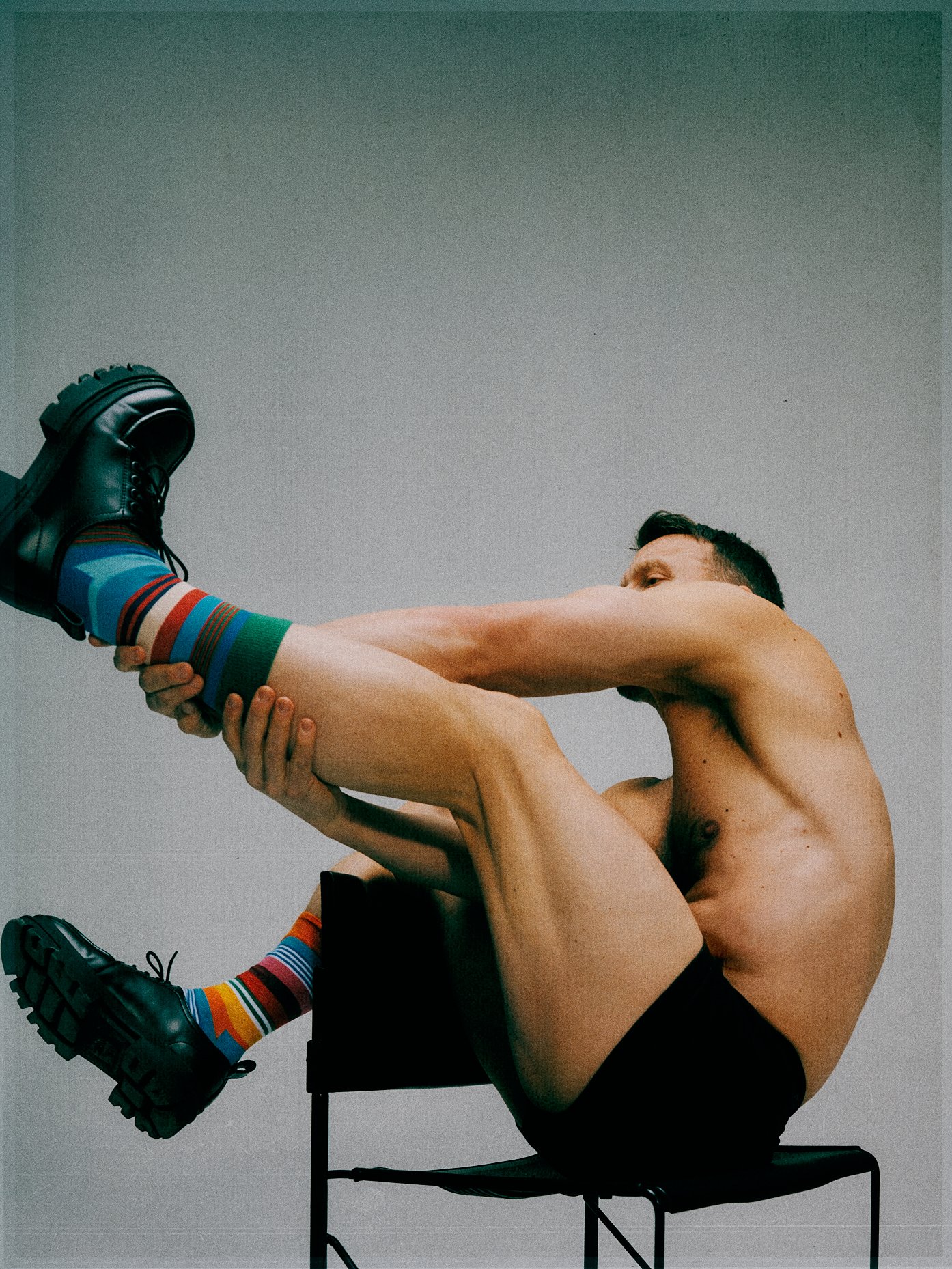 Shirtless man curled sideways on a metal stool while holding one leg across his body.