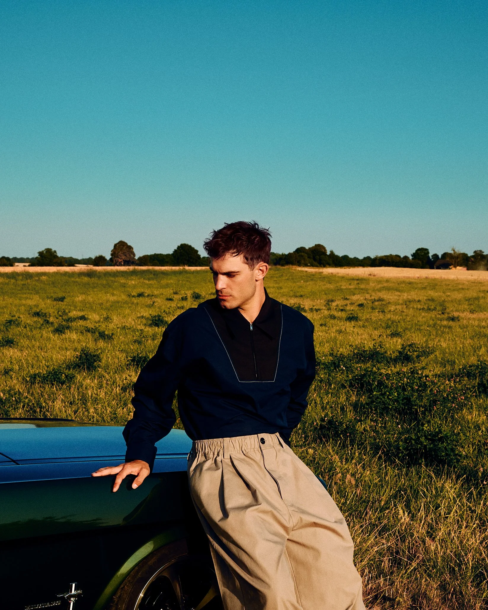 Man in a navy top and pleated beige trousers leaning against a convertible in a field.