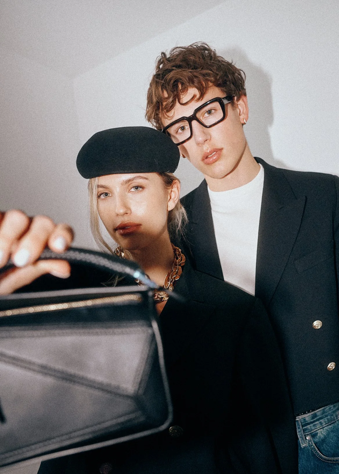 Woman in a black beret holding a dark handbag towards the camera beside a man in glasses.