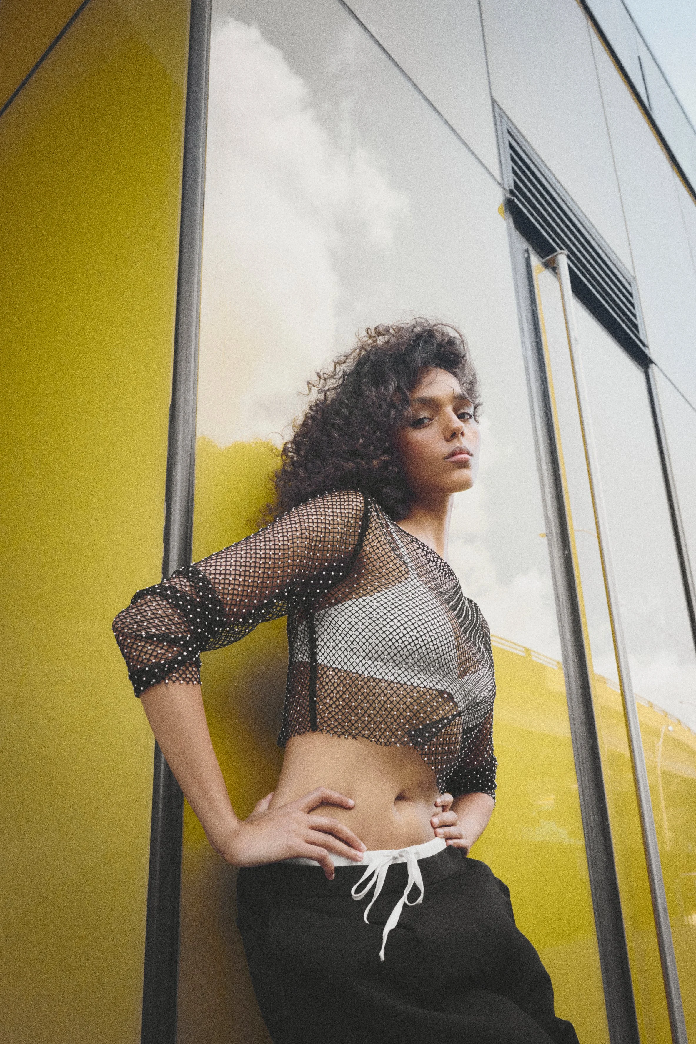 Woman in a sheer black top leaning against a yellow-and-white wall.