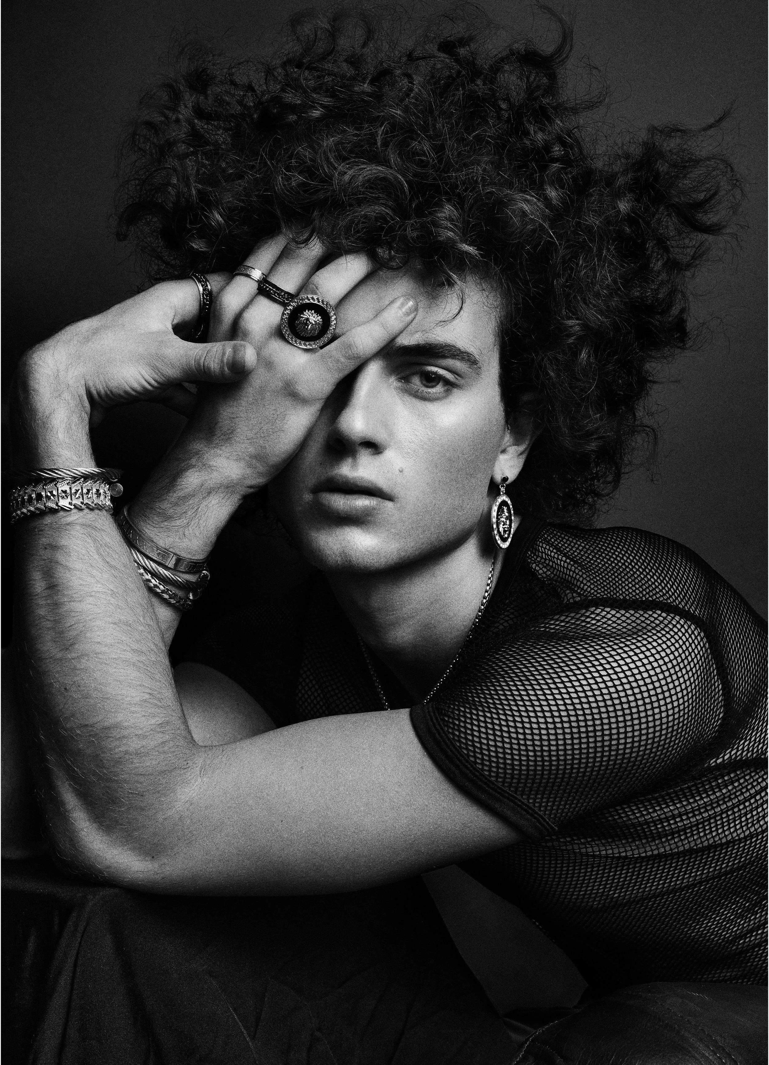 Black-and-white portrait of a curly-haired model wearing rings and a sheer top, with one hand across the forehead.