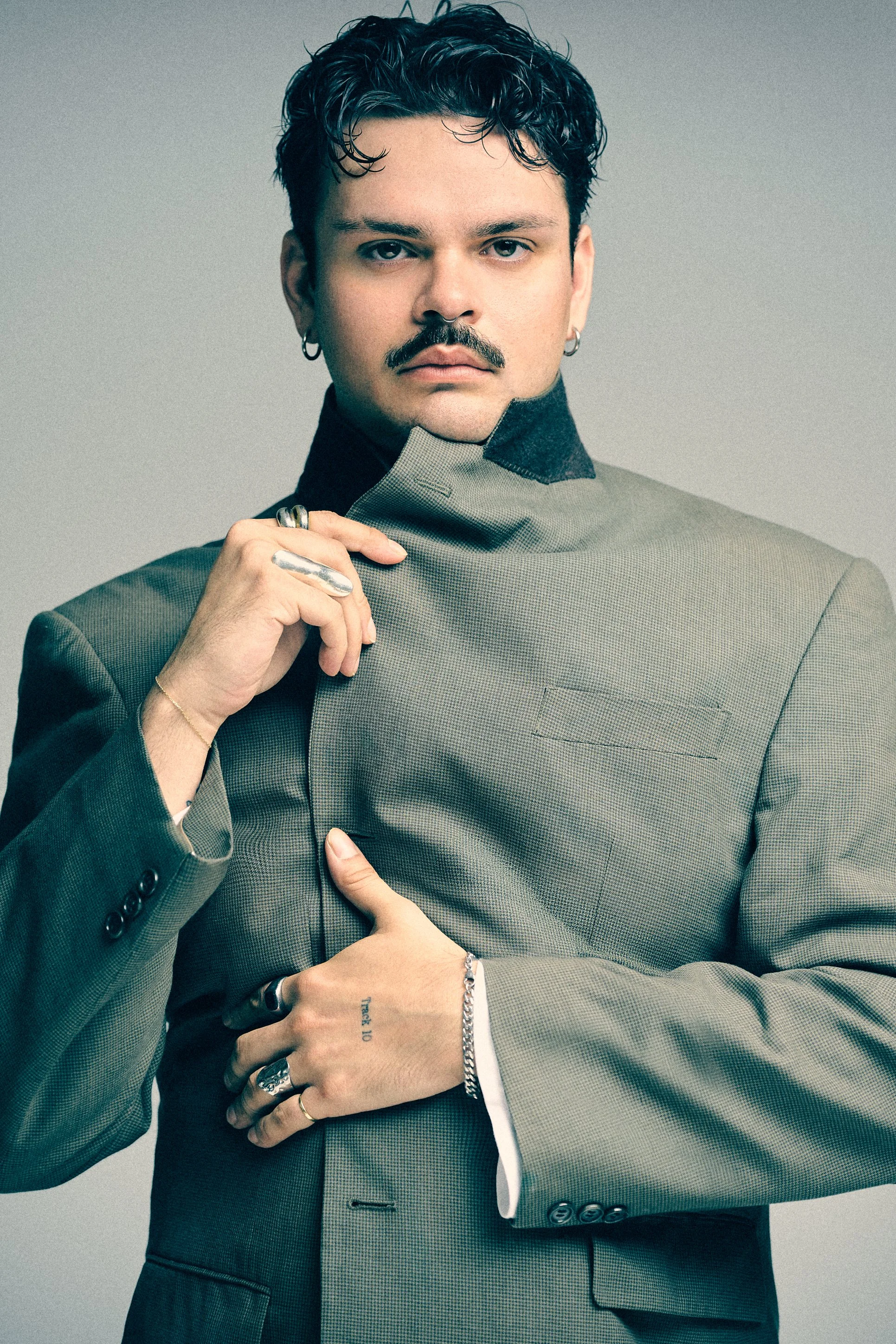 Man with a moustache and curled hair wearing a high-collared grey jacket and silver rings.