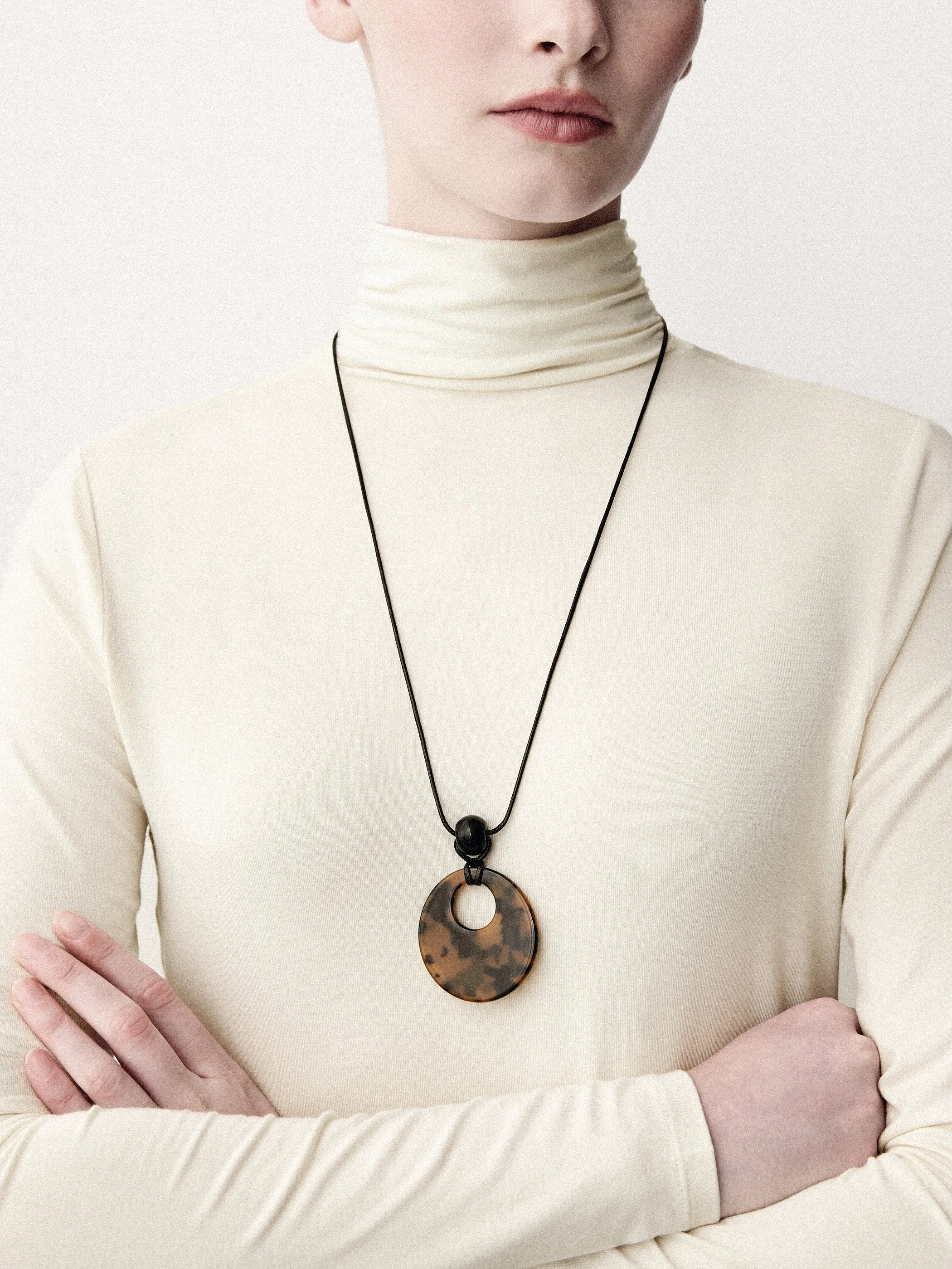 Detail of a cream turtleneck with a large circular pendant and crossed arms.