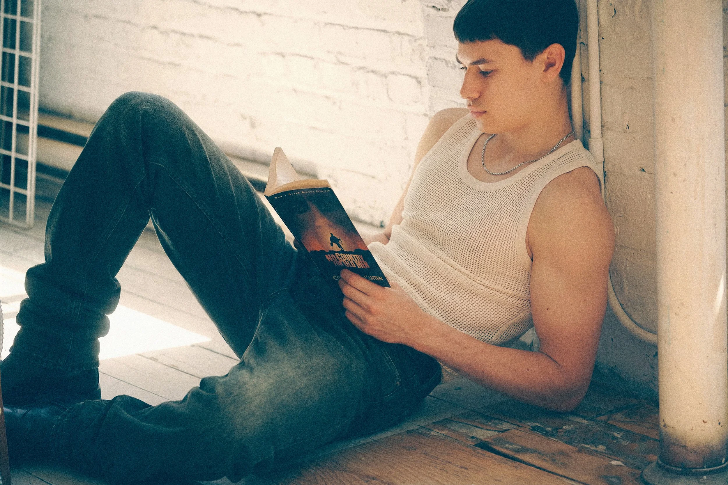 Young man in a white tank top and jeans reclining beside a window while reading a book.
