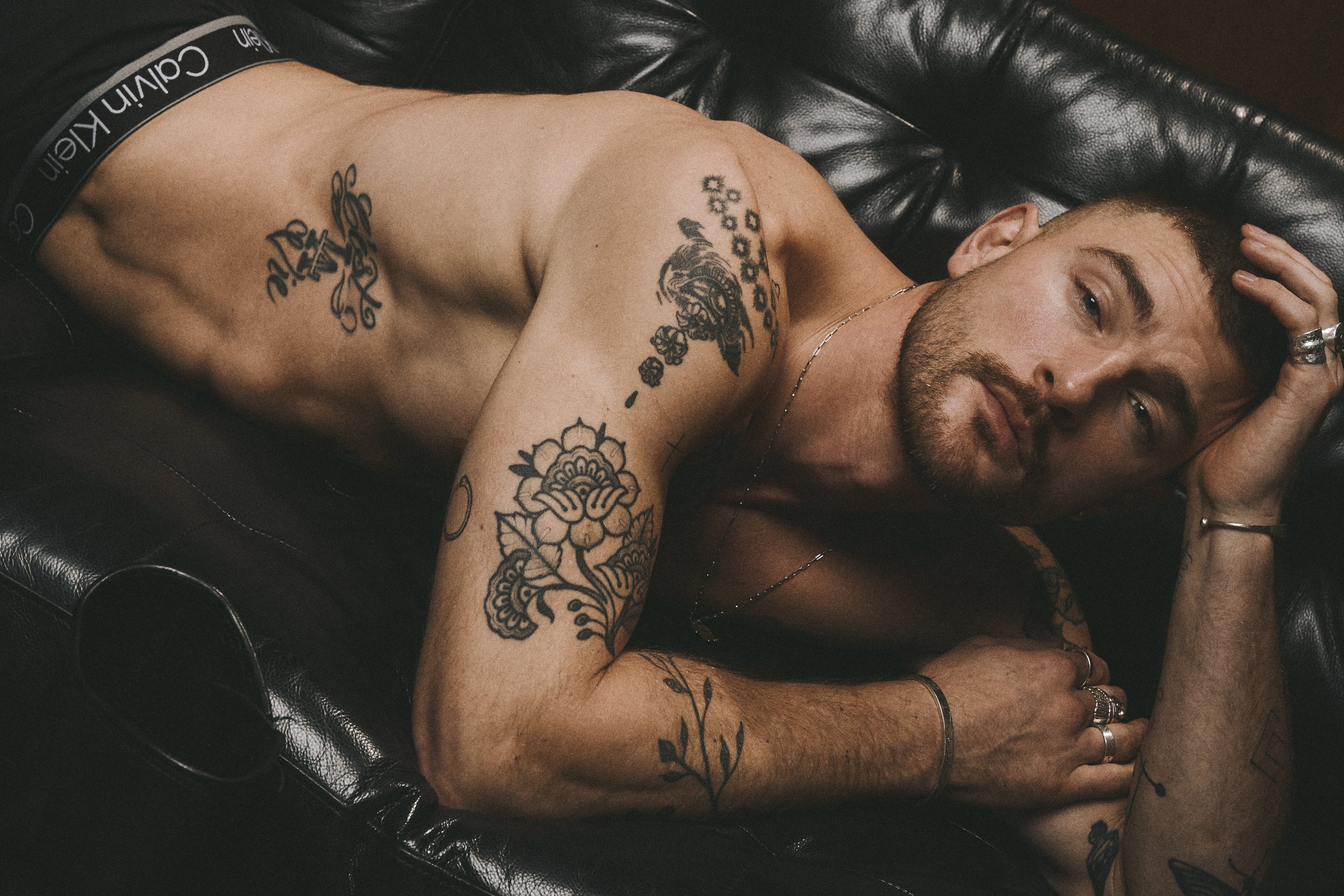 Close view of a shirtless tattooed man lying on a leather sofa with his hands together.
