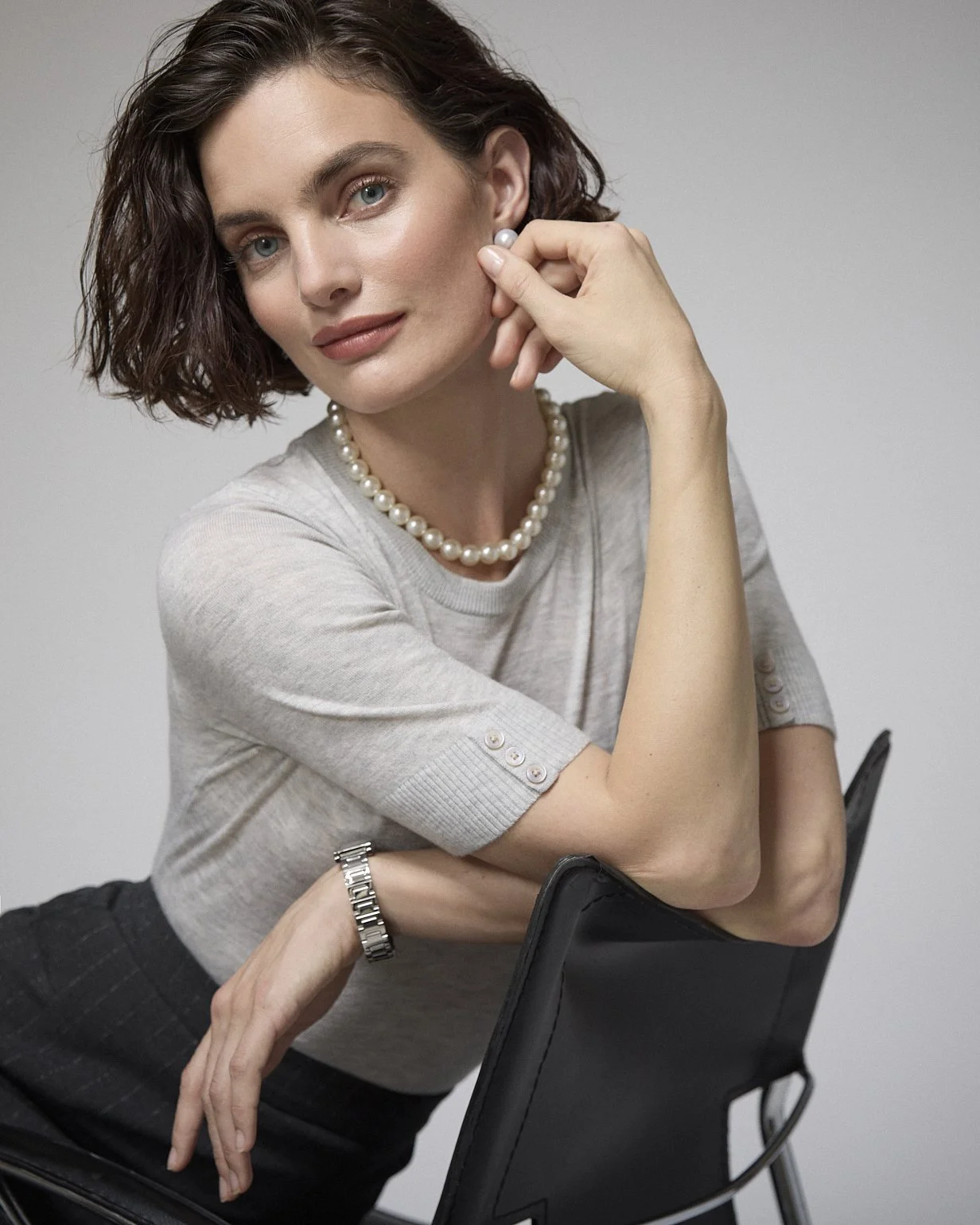 Close portrait of a woman in a grey knit top and pearl necklace seated on an office chair.