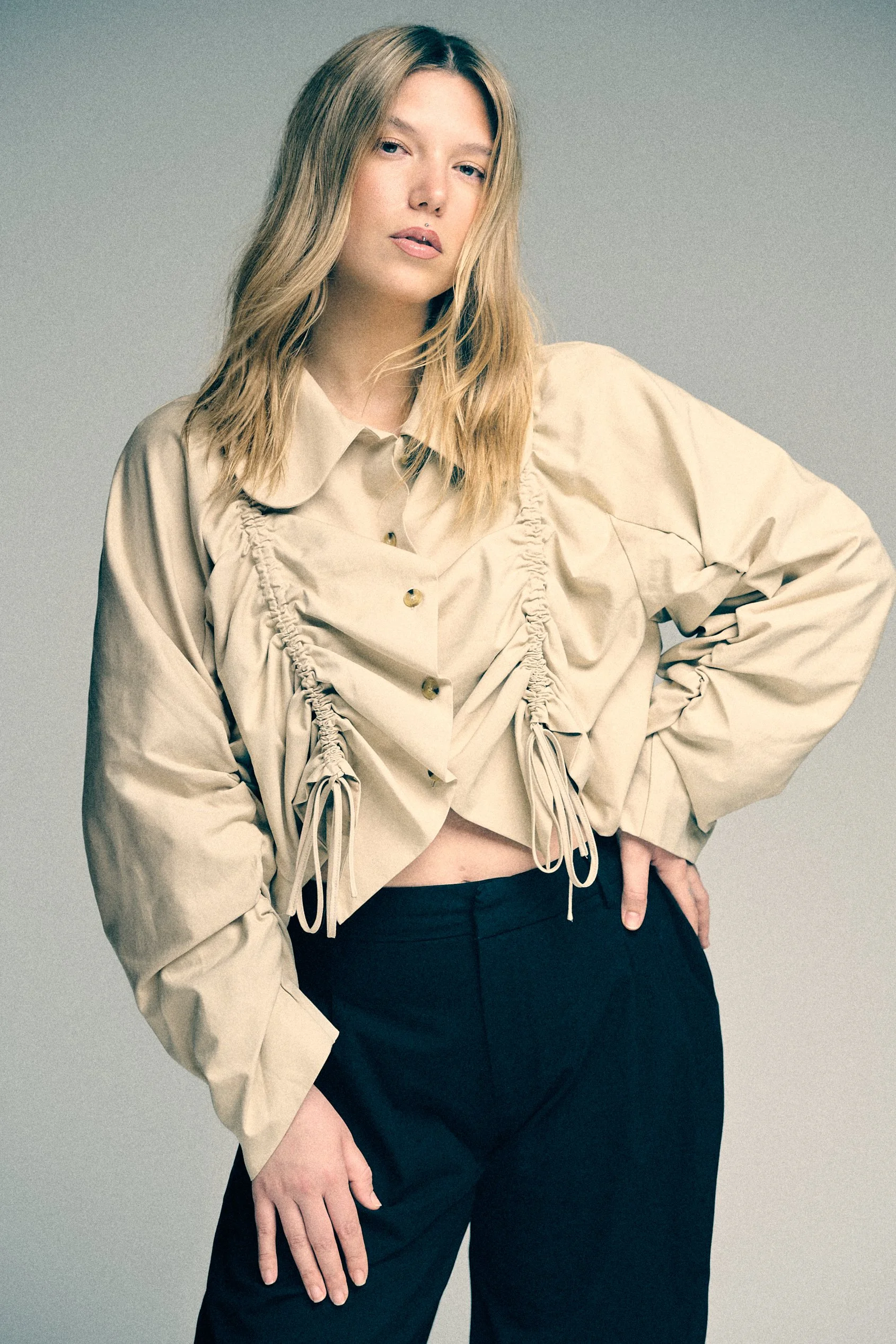 Woman in a cream gathered blouse and dark trousers, with one hand on her hip.