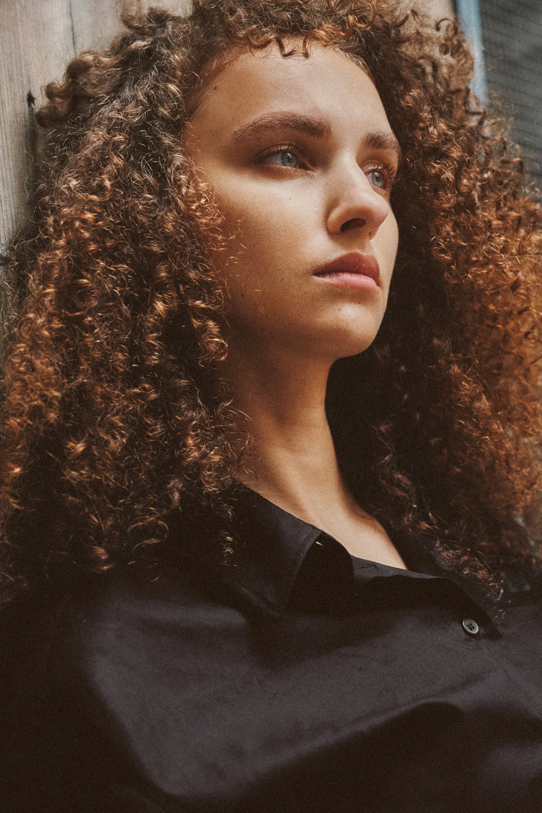 Close-up fashion portrait of a curly-haired woman in a black shirt gazing off-camera, with warm light and a grainy film texture.
