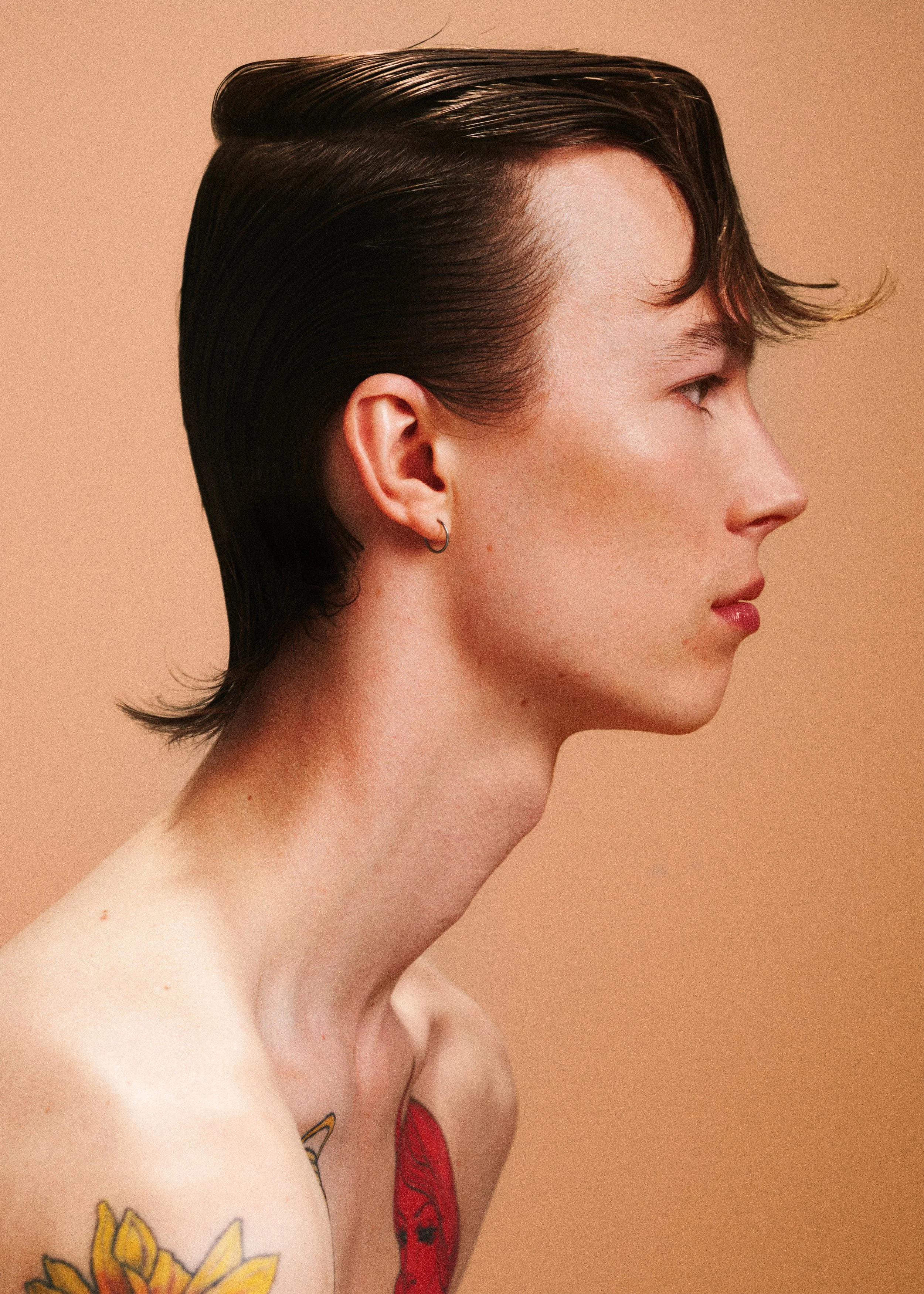 Side-profile portrait of a tattooed man with slicked dark hair against a peach background.