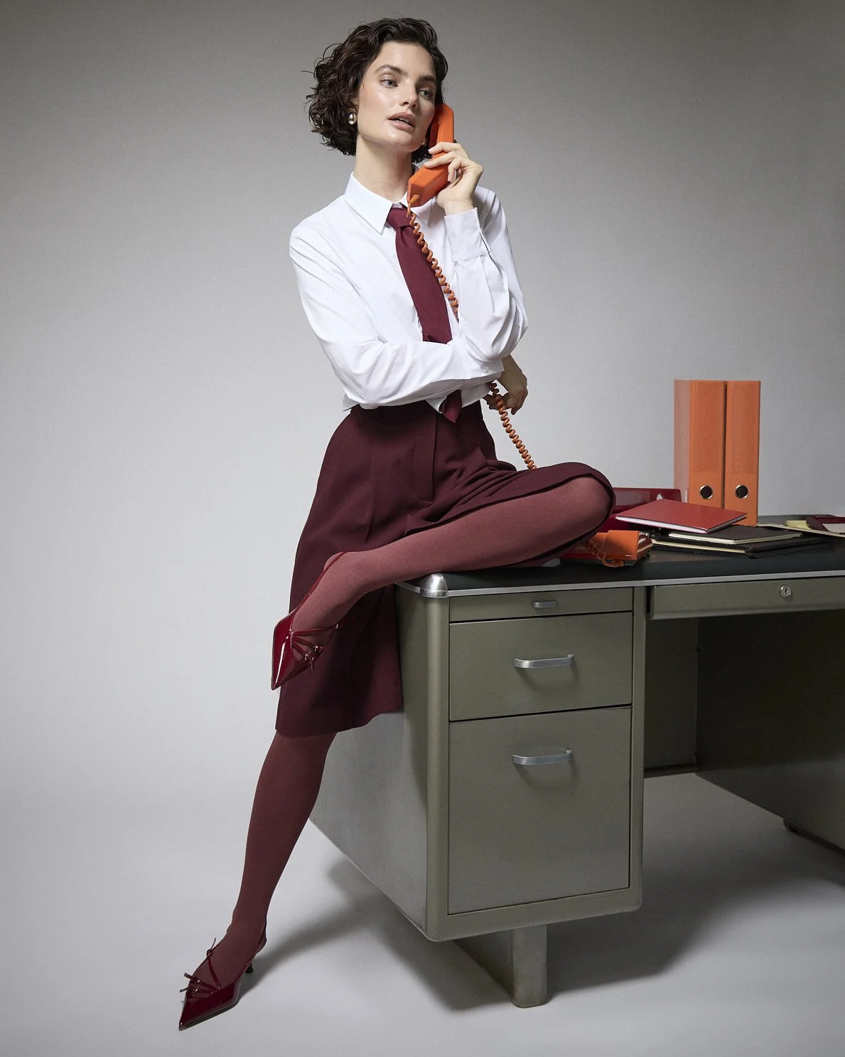 Woman perched on an office desk in a white shirt, burgundy tie and trousers, holding an orange telephone.