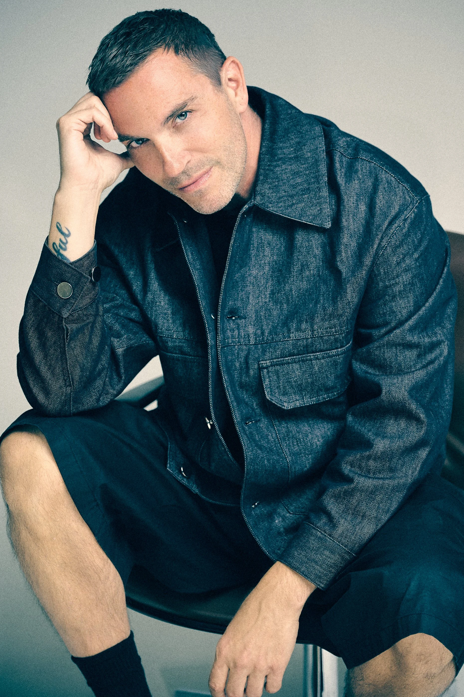 Man in a dark denim shirt and shorts seated with his head resting on one hand.