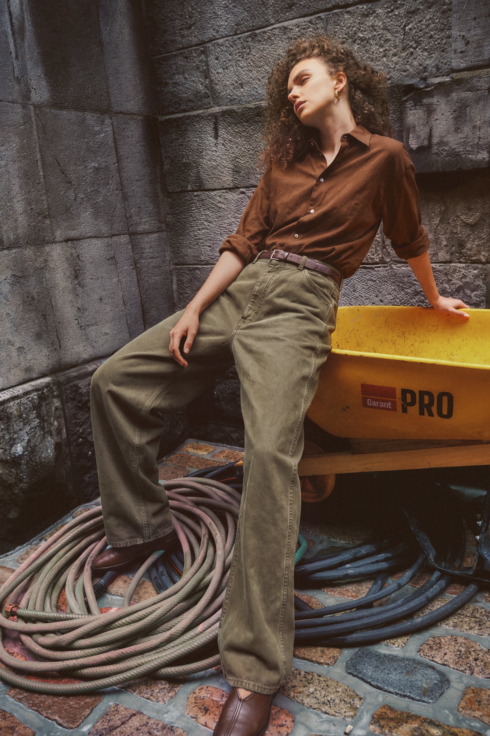 Curly-haired fashion model in a brown shirt and olive trousers leaning against a yellow wheelbarrow in a stone courtyard.
