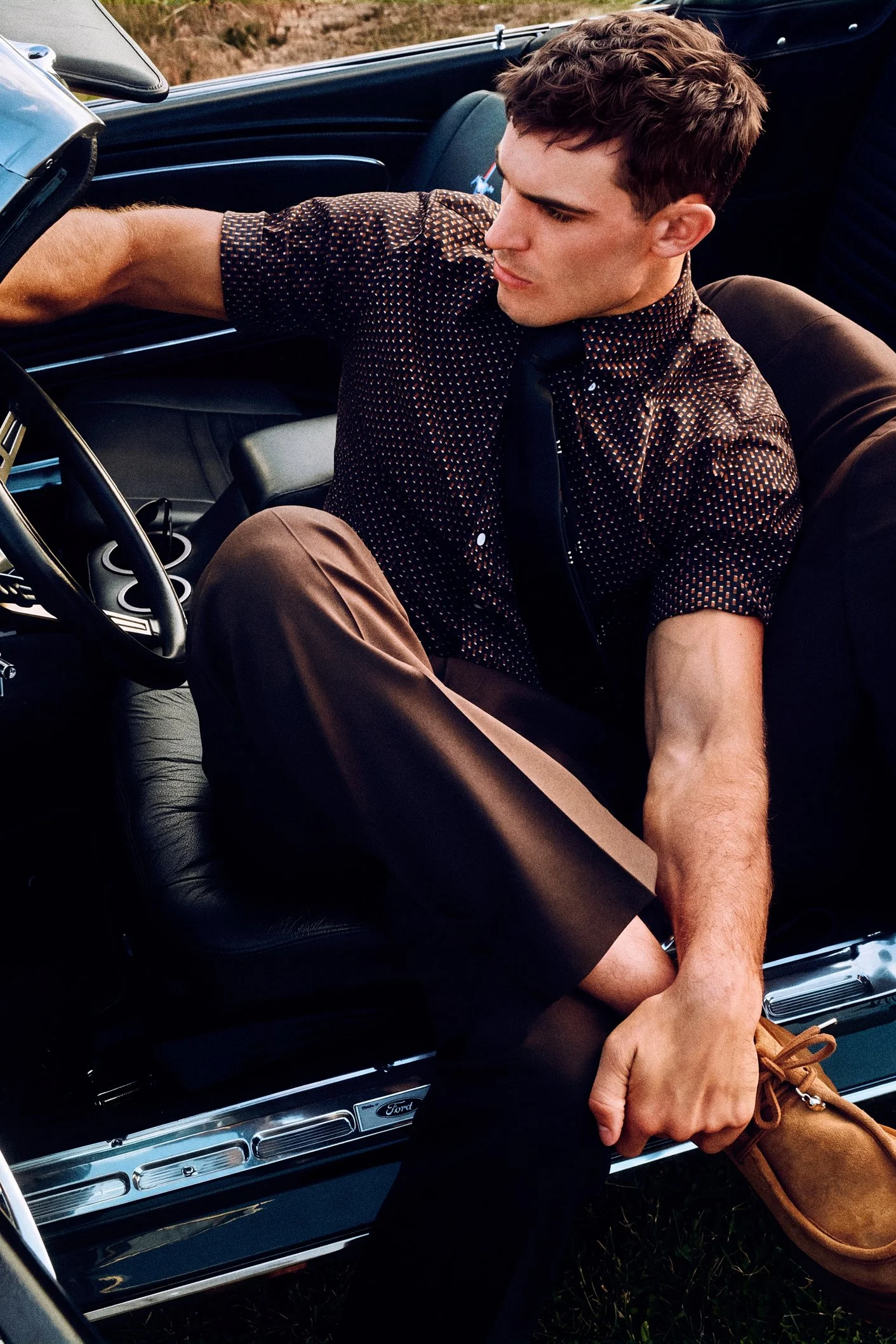 Man in a patterned dark shirt seated sideways in a convertible, with one hand on the door.