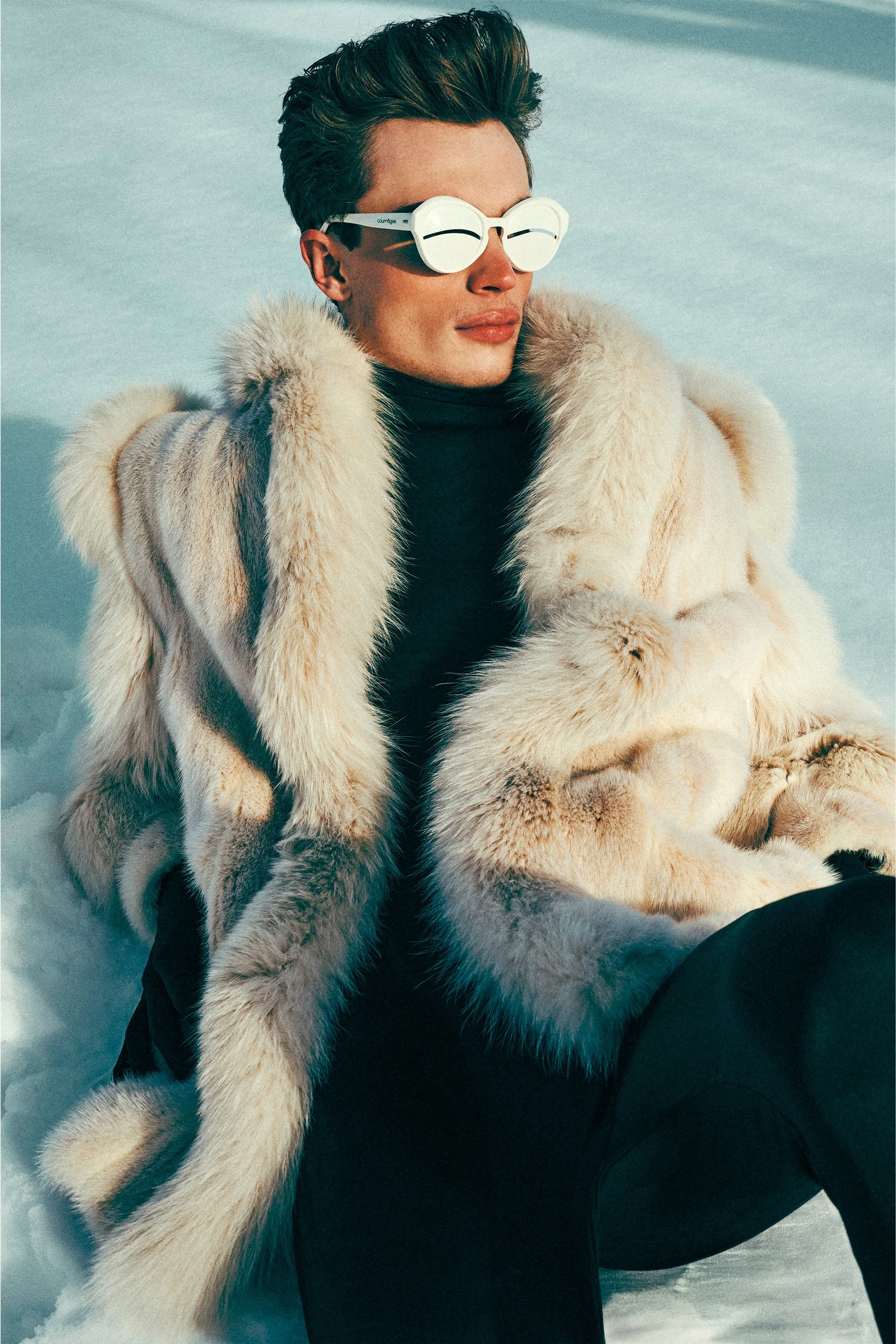 Man in Inuit inspired sunglasses and a pale fur-textured coat seated in snow.