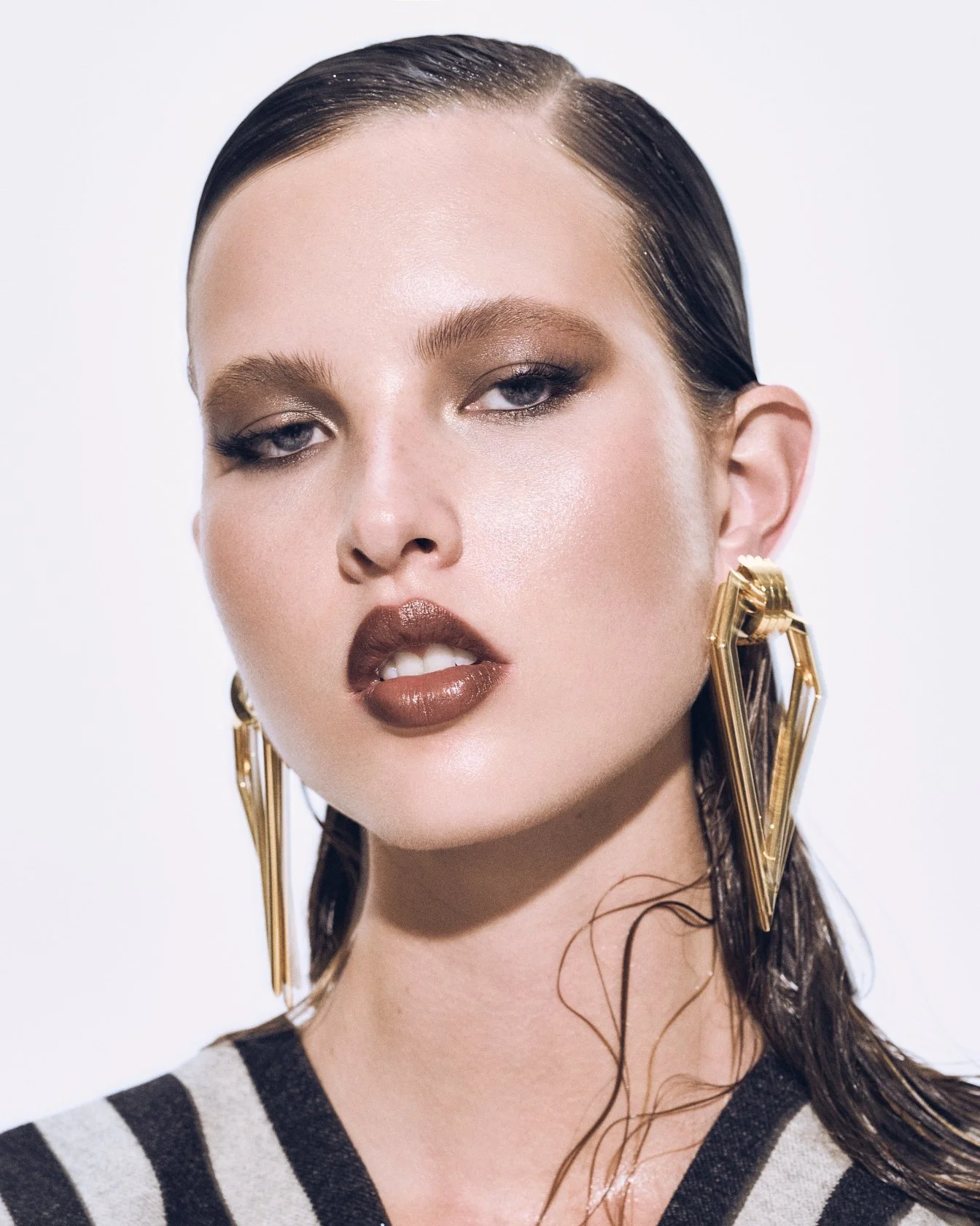 Beauty close-up of a woman with slicked hair and dark lipstick, wearing large gold earrings and a graphic black-and-white jacket.