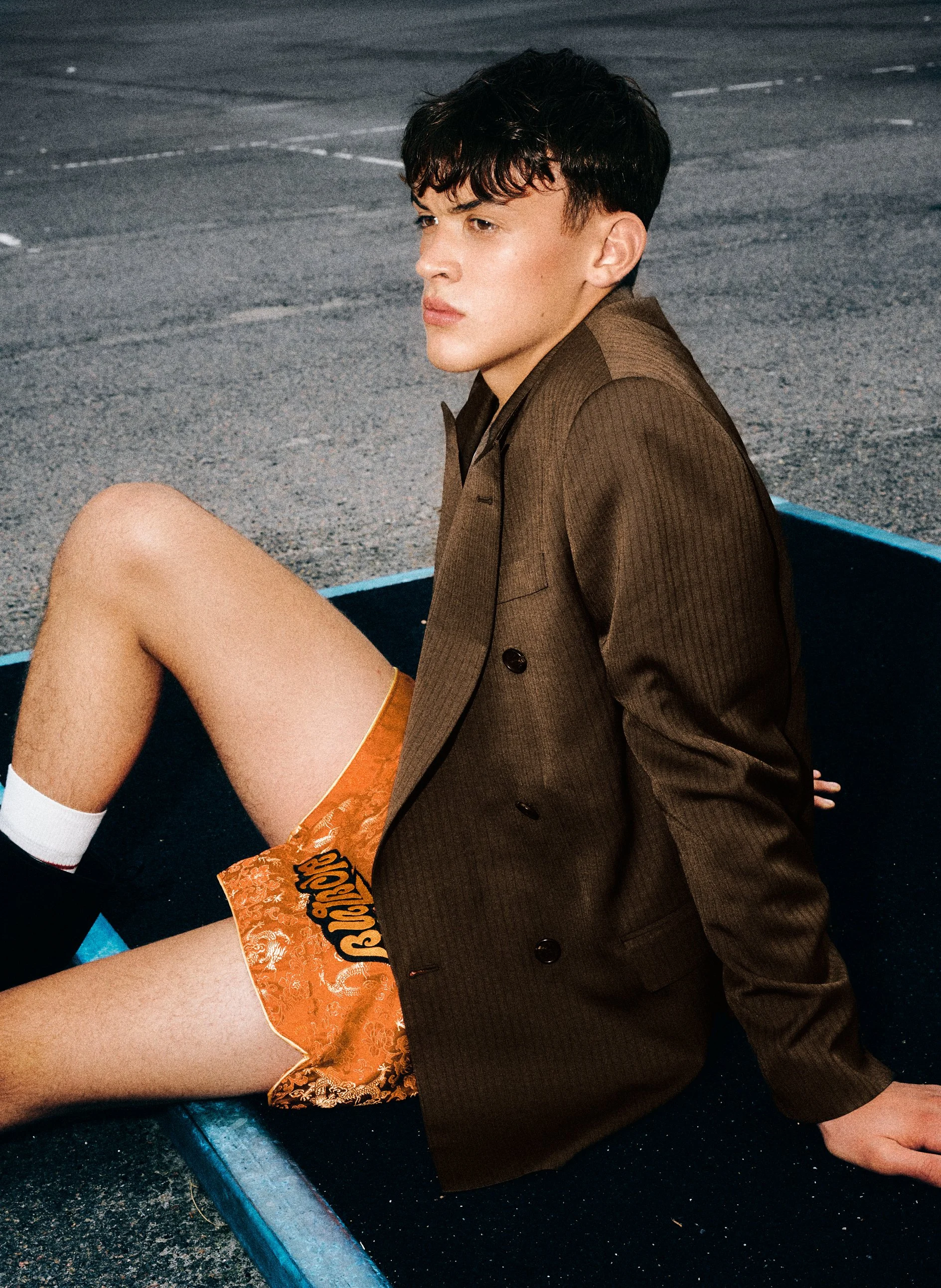 Man in a brown jacket and orange patterned shorts seated on outdoor exercise equipment.