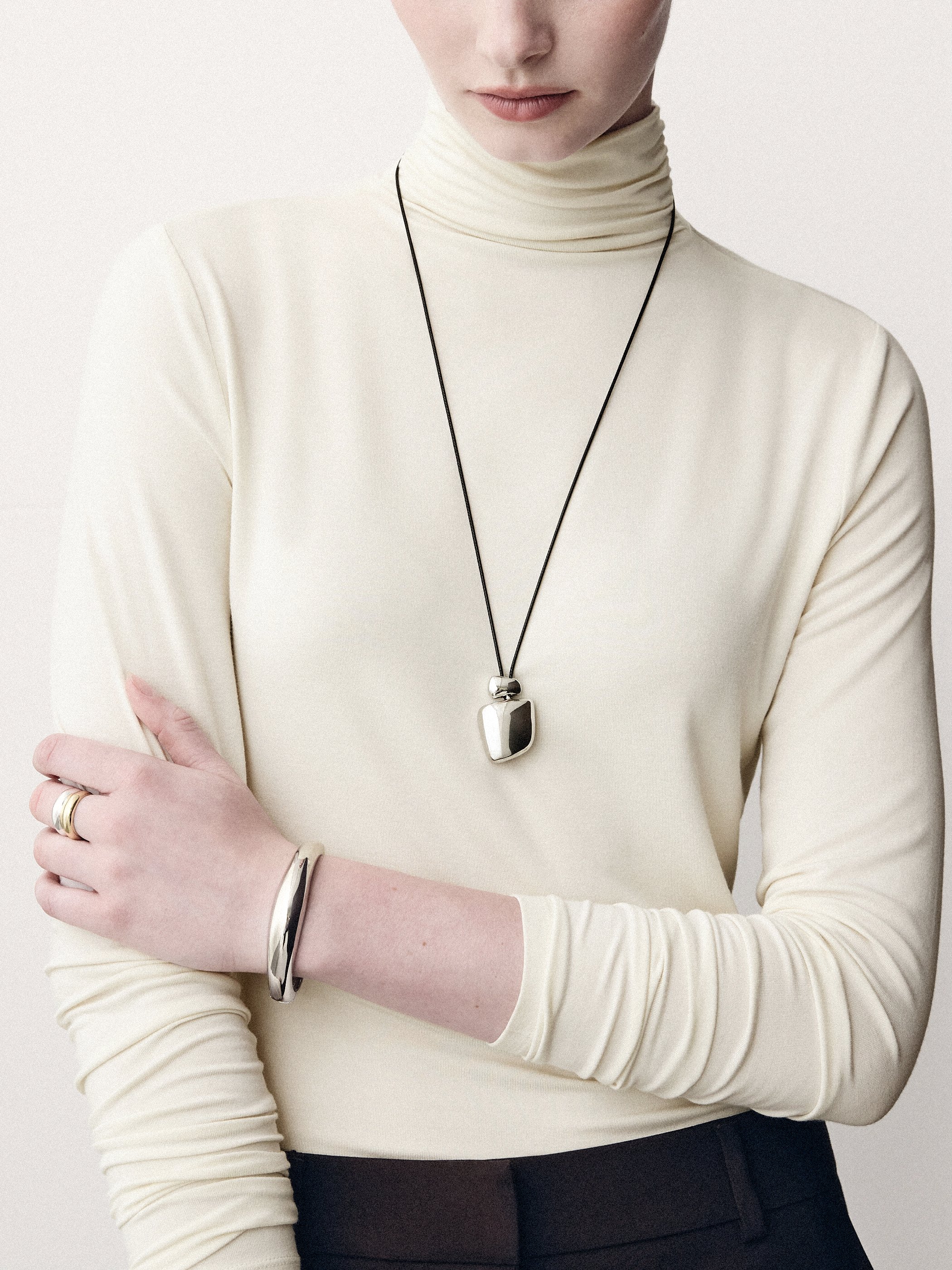 Detail of a cream turtleneck, silver pendant necklace and crossed arms with bracelets.