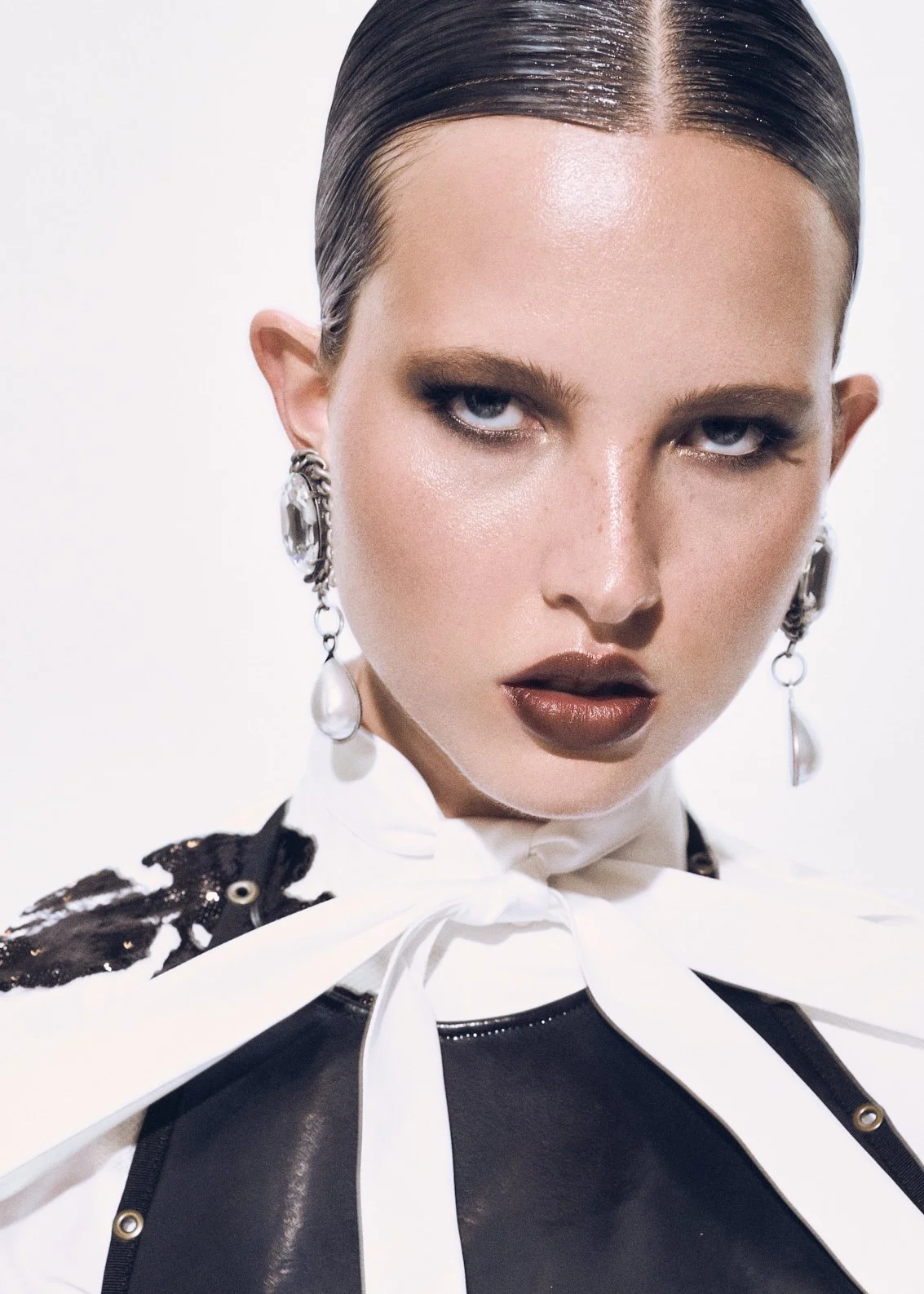 Beauty close-up of a woman with slicked hair, dark lipstick, dangling earrings and a large white bow.