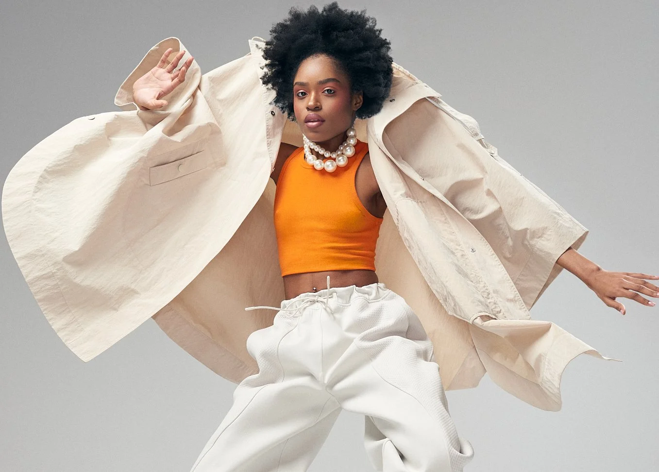 Woman raising her arms in an orange top, white trousers and billowing beige coat.