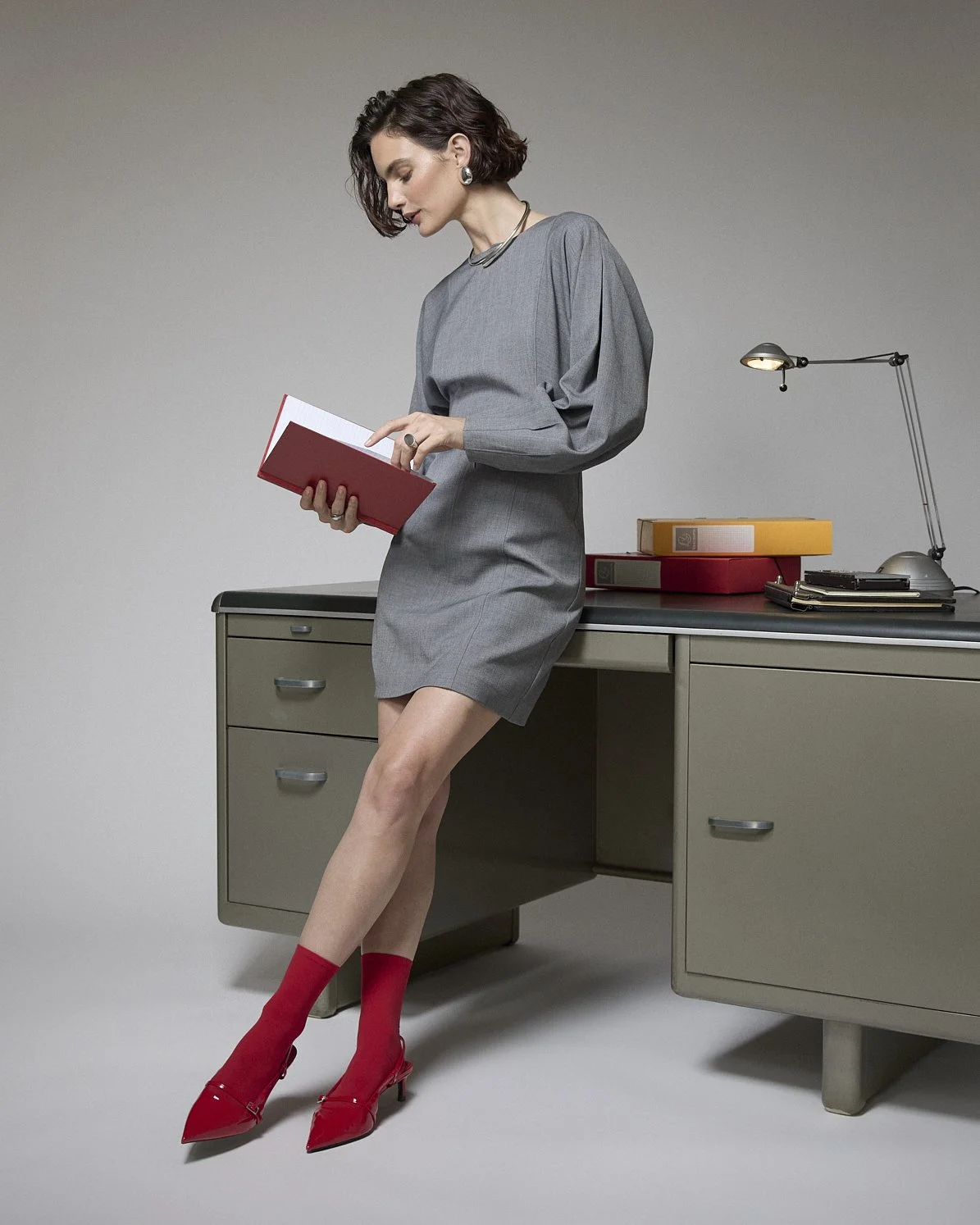 Woman in a grey dress and red boots leaning against a desk while reading a small book.