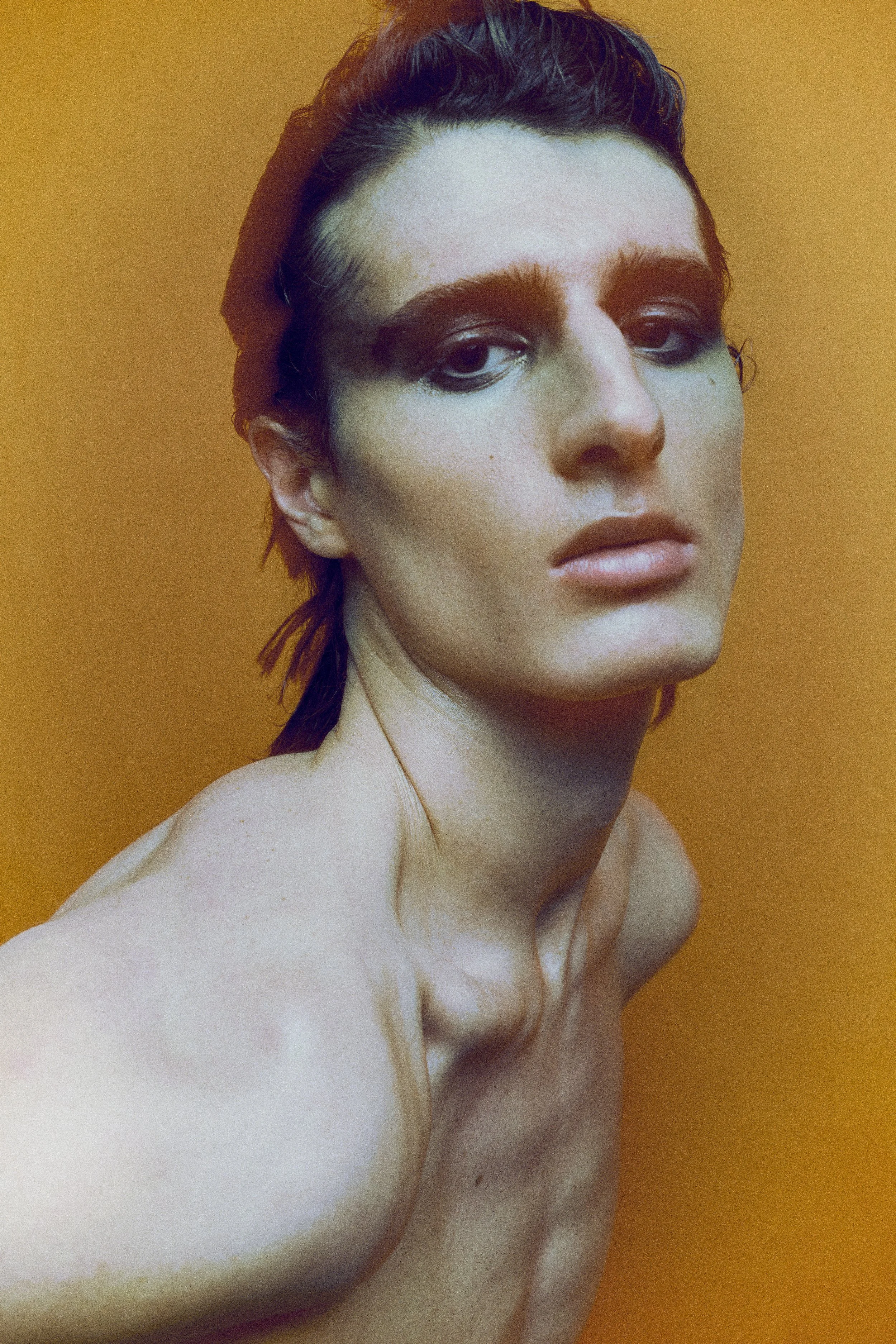 Close portrait of a bare-shouldered model with purple hair and pale makeup against a yellow background.