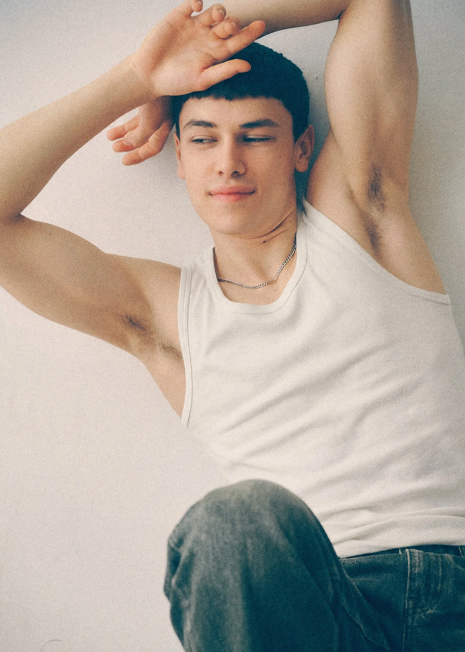 Young man in a white tank top seated with both arms raised above his head.