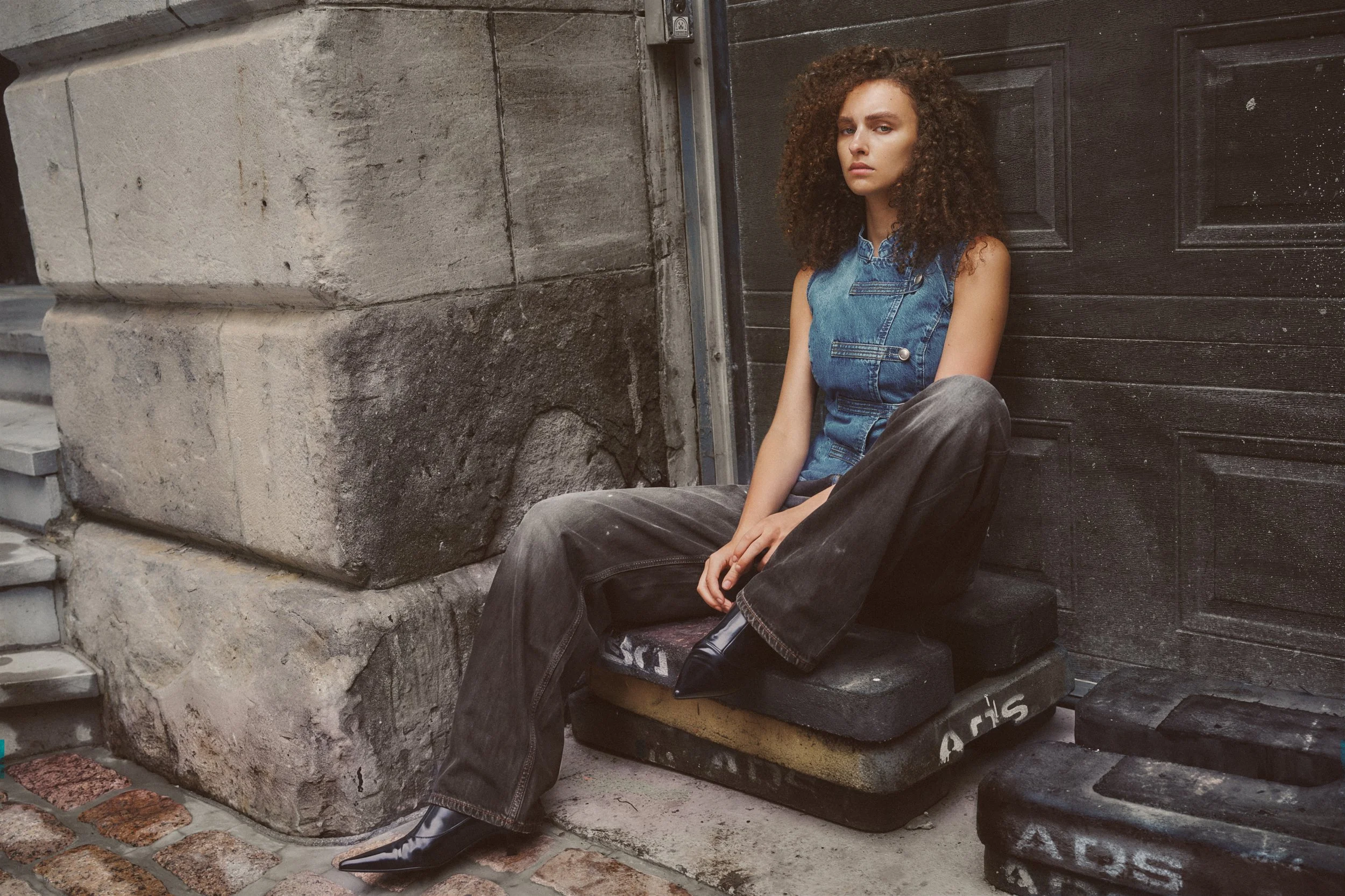 Seated fashion model in a blue denim top, oversized grey jeans and pointed black boots against a weathered stone wall and dark doorway.