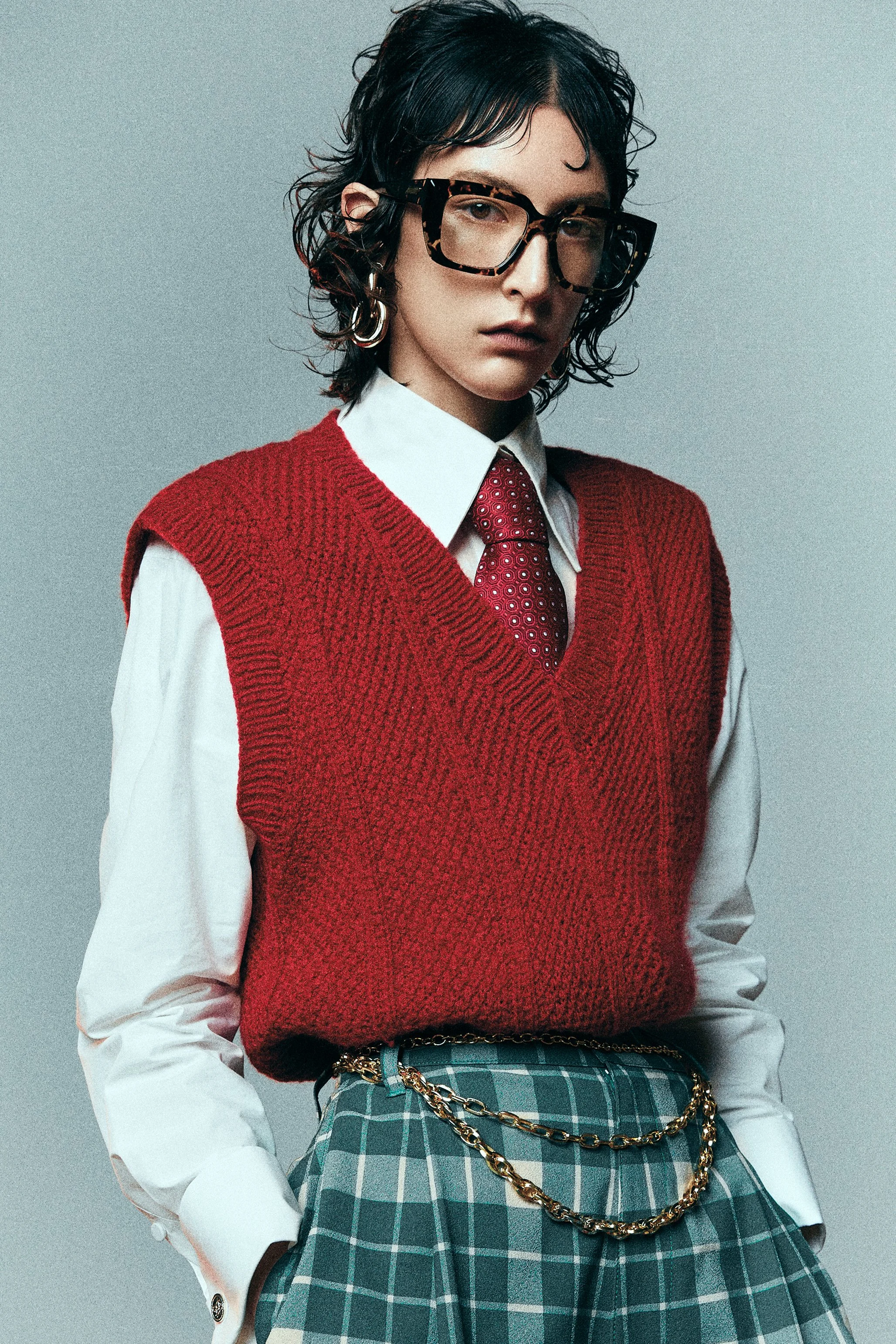 Woman wearing glasses, a red knitted vest, white shirt and tie with a plaid skirt and chain belt.