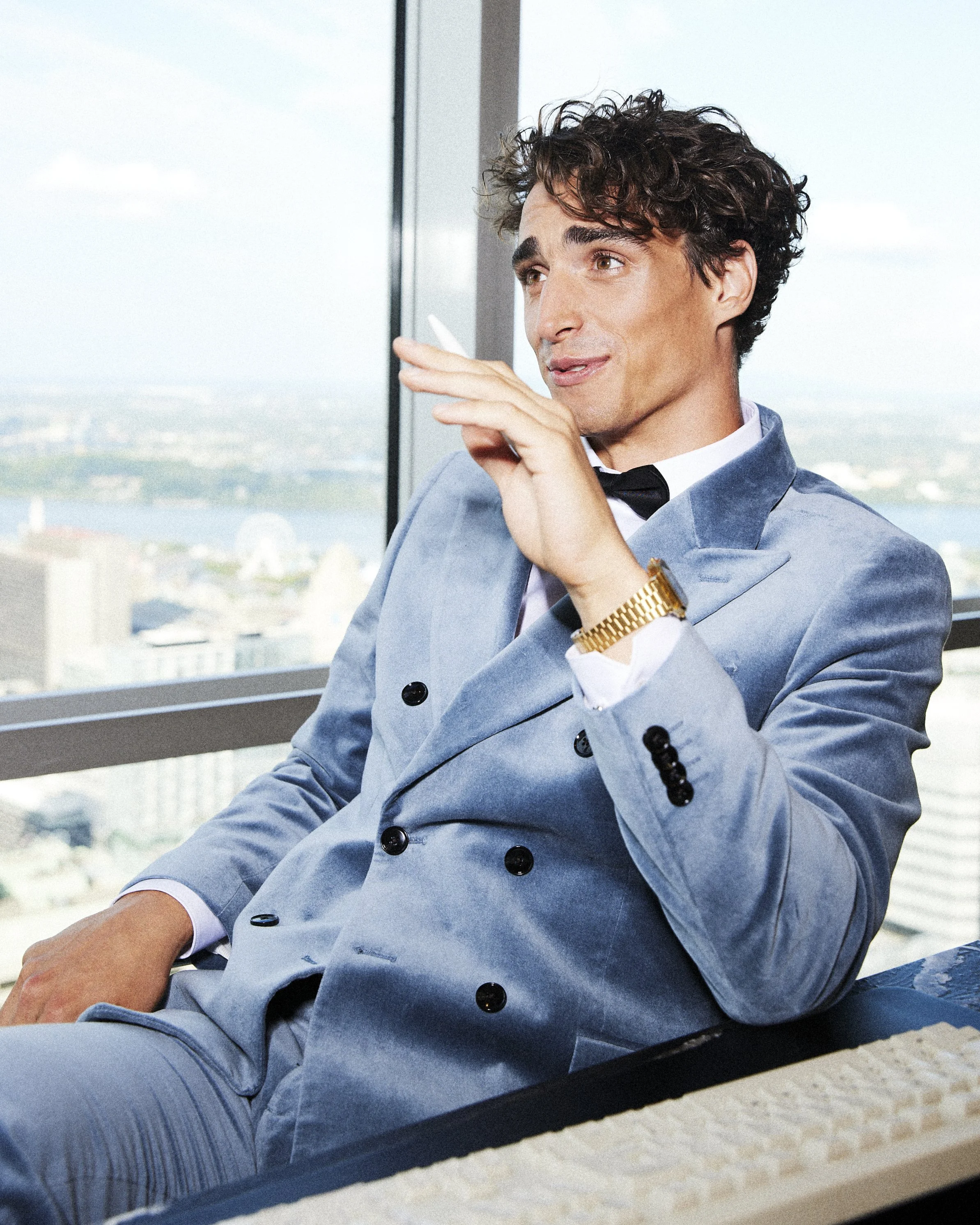 Man seated by a large window in a pale blue tuxedo with one hand raised.