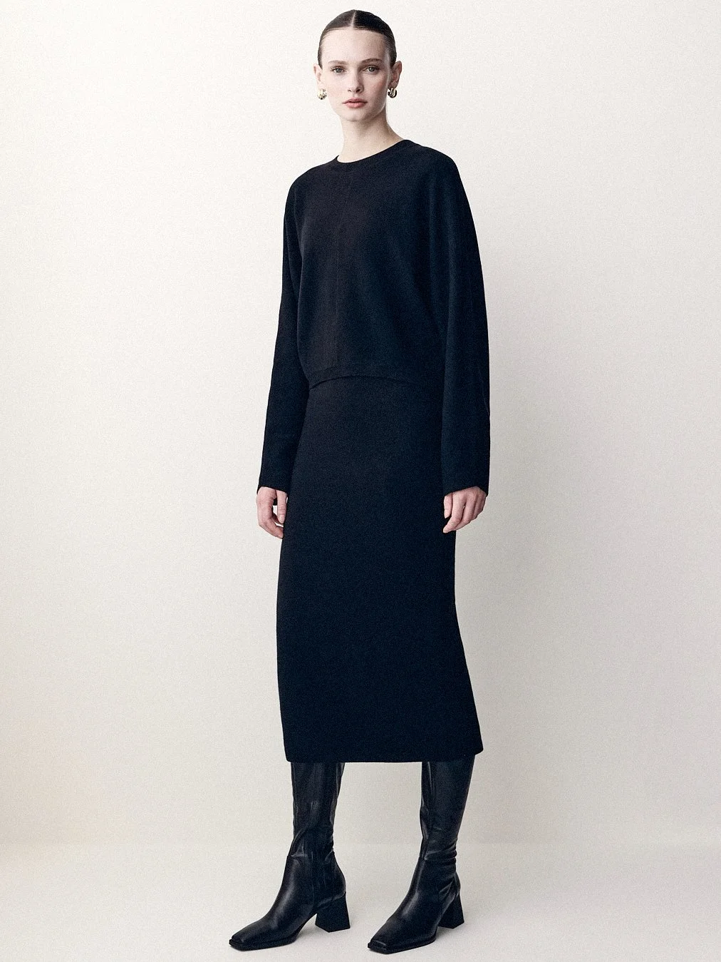 Woman in a long black knit dress and knee-high boots standing in the studio.
