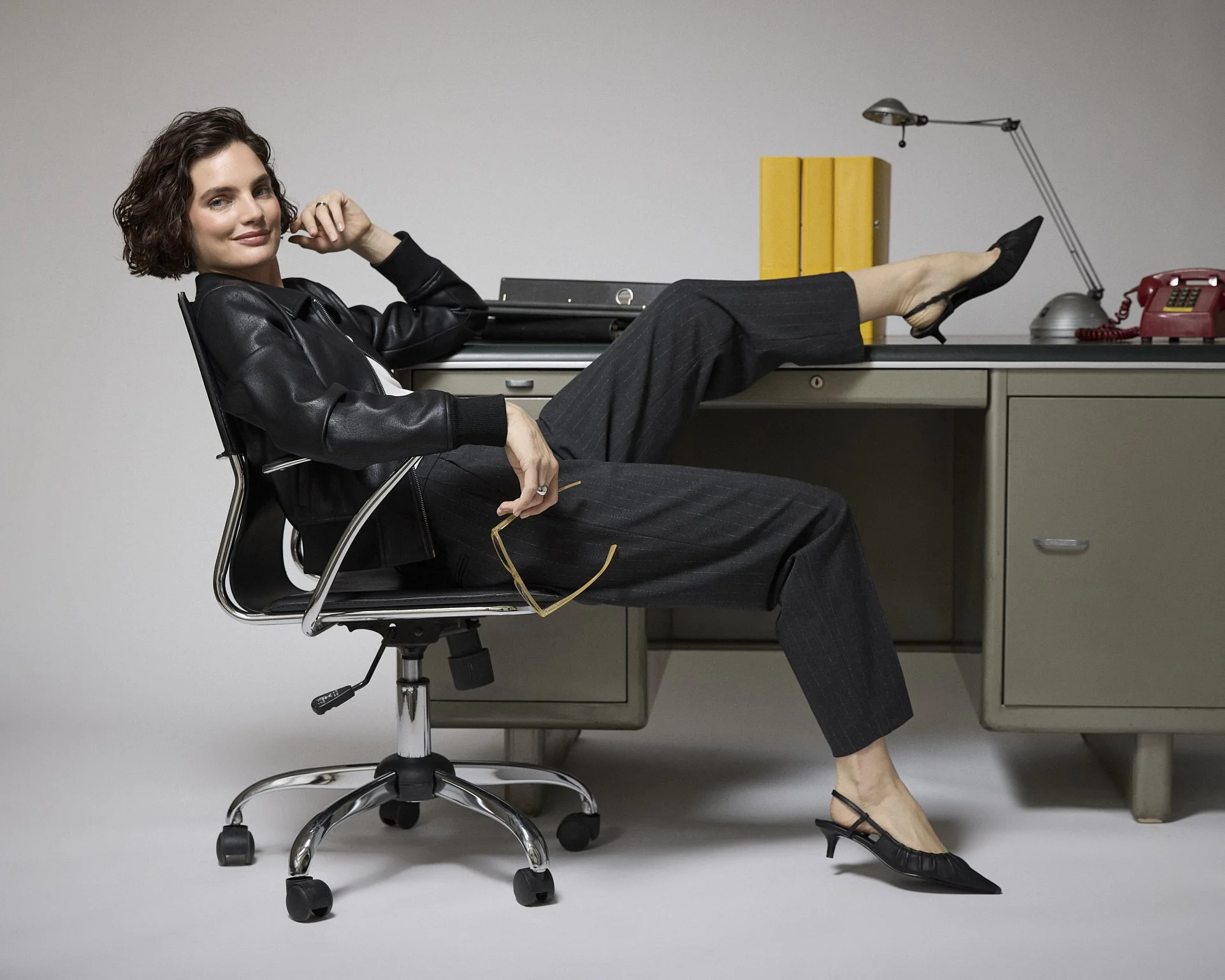 Woman in a black outfit reclining on an office chair with her feet resting on a desk.