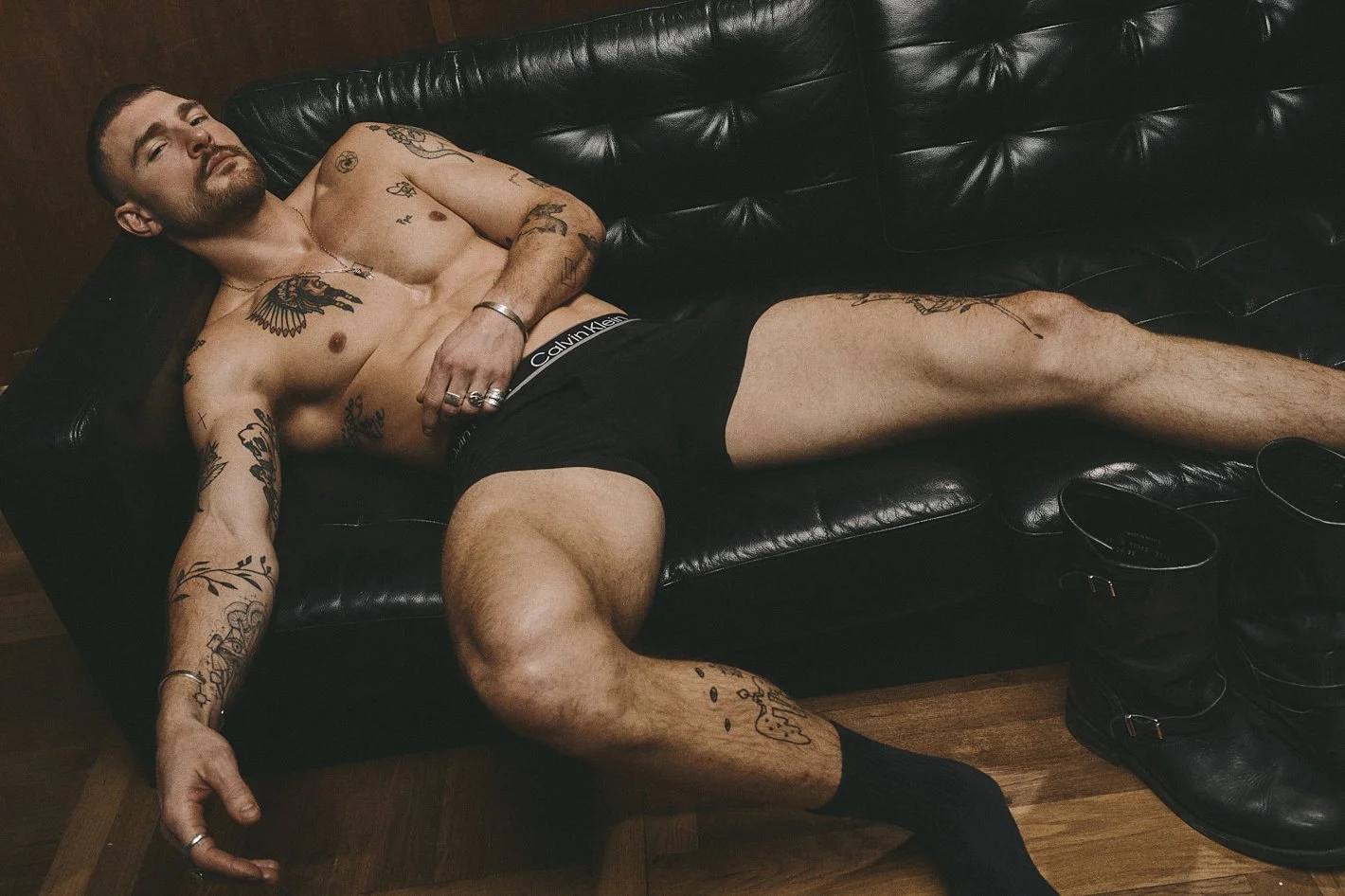 Shirtless tattooed man reclining on a black leather sofa in dark shorts.