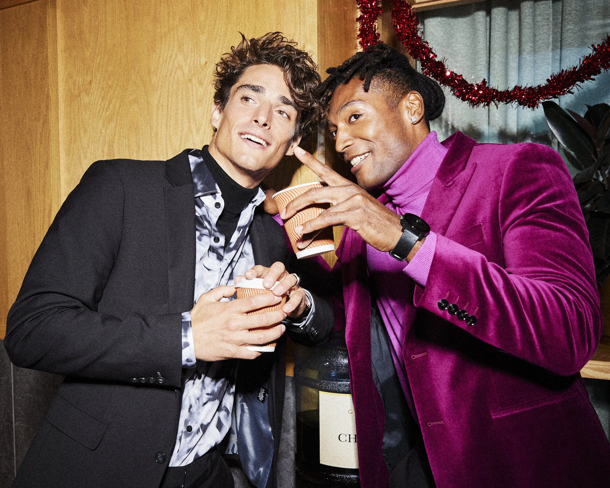 Two men laughing at a holiday party in black and burgundy tailoring while holding drinks.