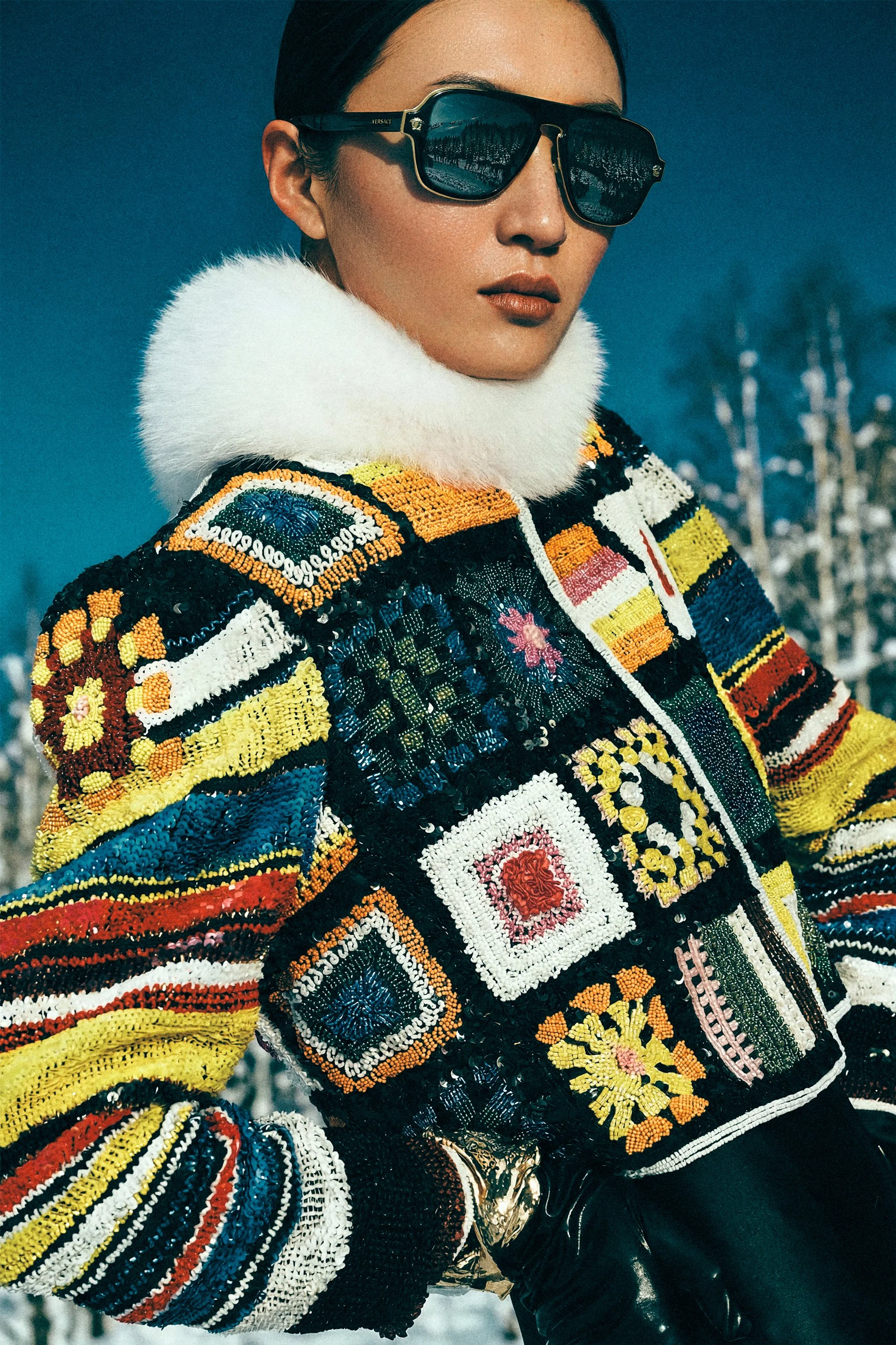 Close portrait of a woman in oversized sunglasses, colourful knitwear and a white fur collar.