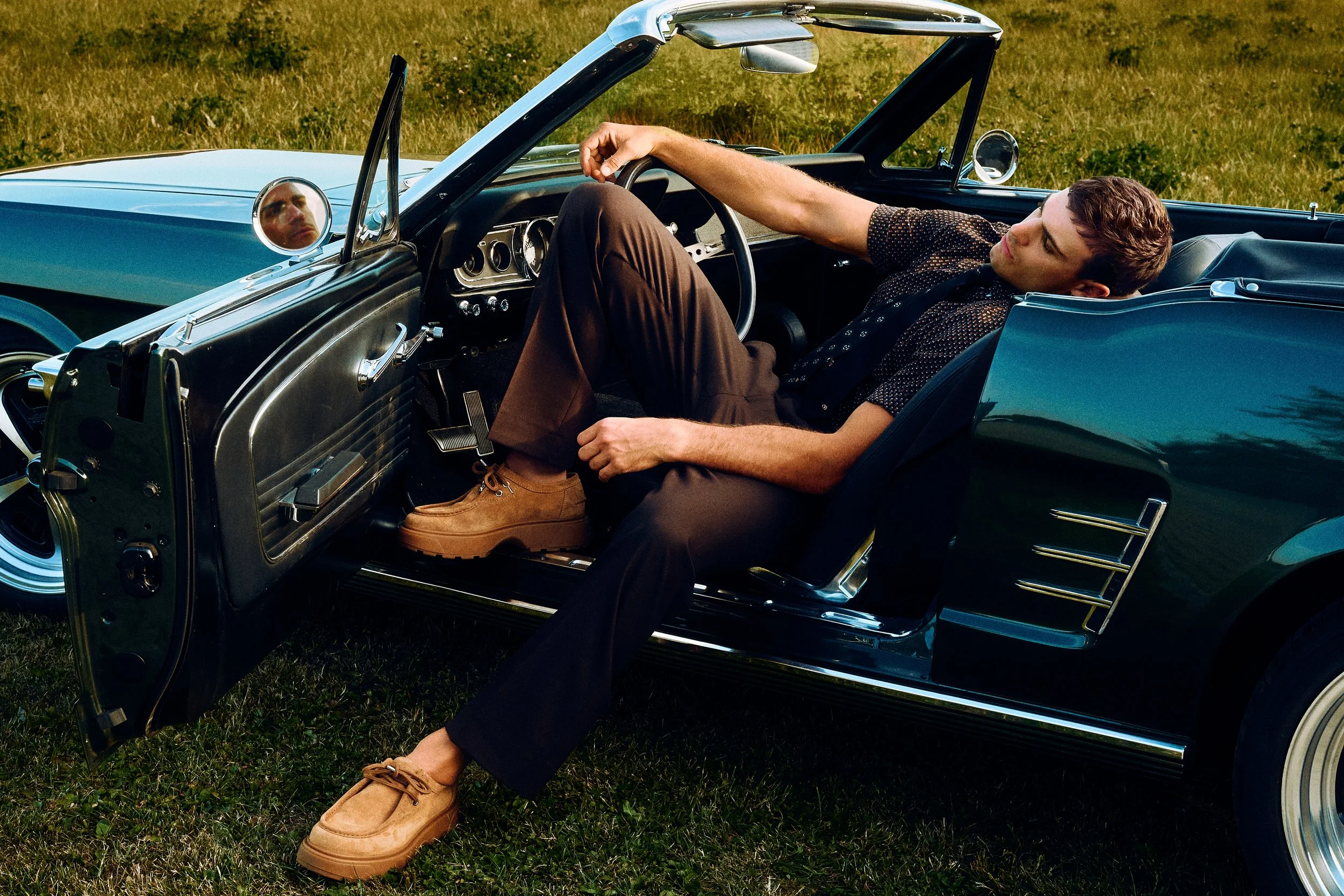 Man reclining in an open convertible, wearing a dark shirt and brown trousers.