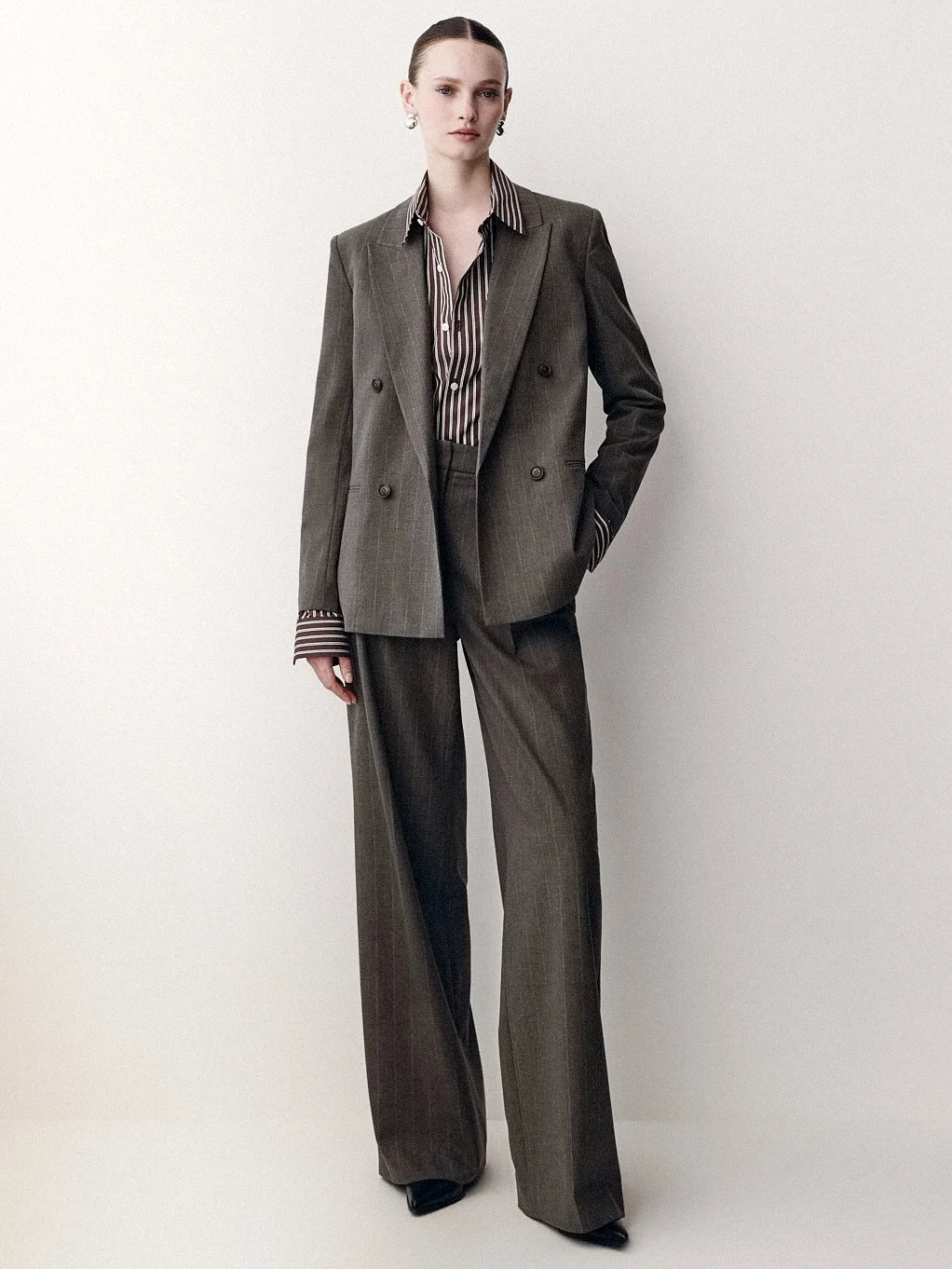 Woman in a brown trouser suit and striped shirt, with one hand in her pocket.