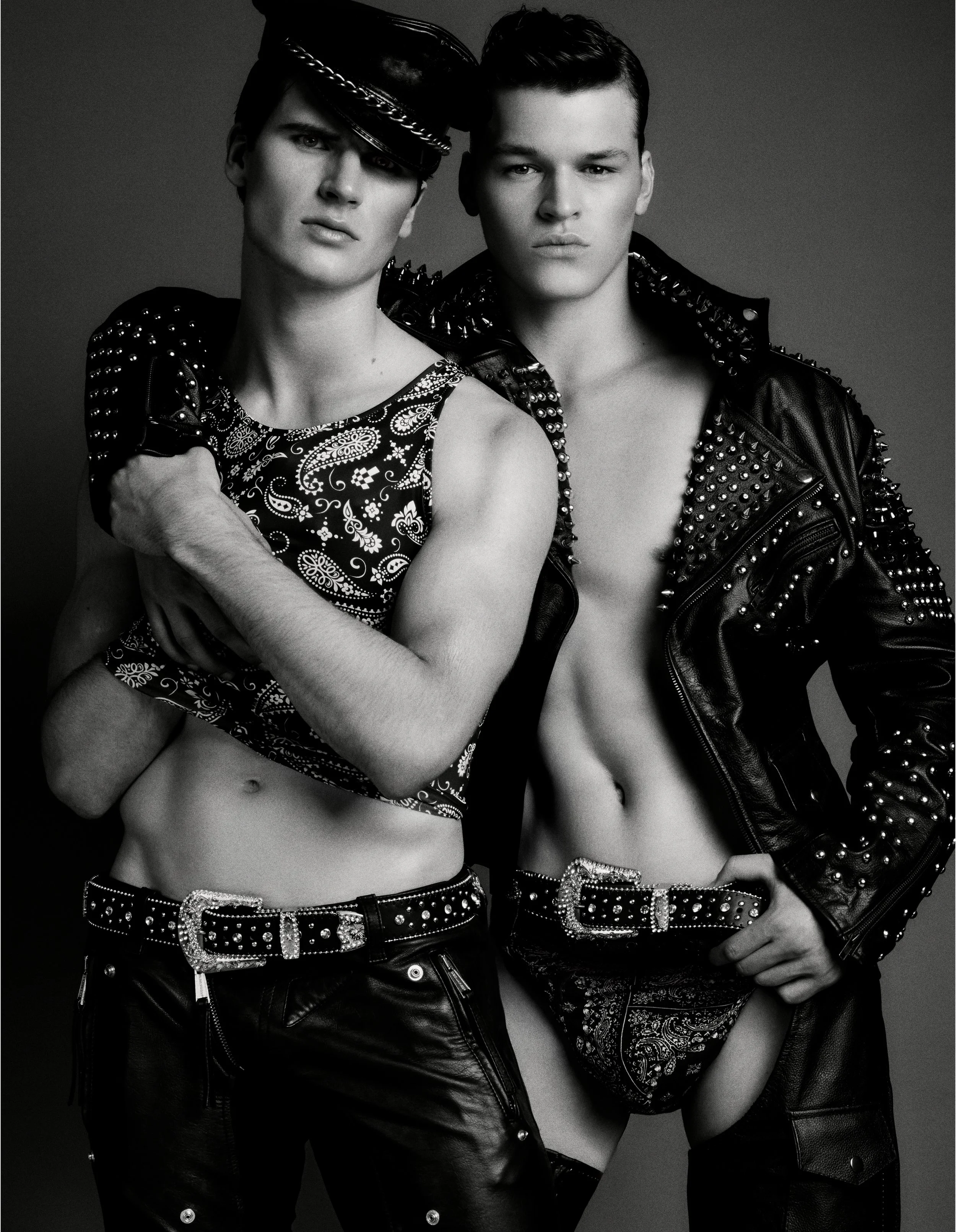 Black-and-white portrait of two men in studded leather and patterned accessories standing close together.