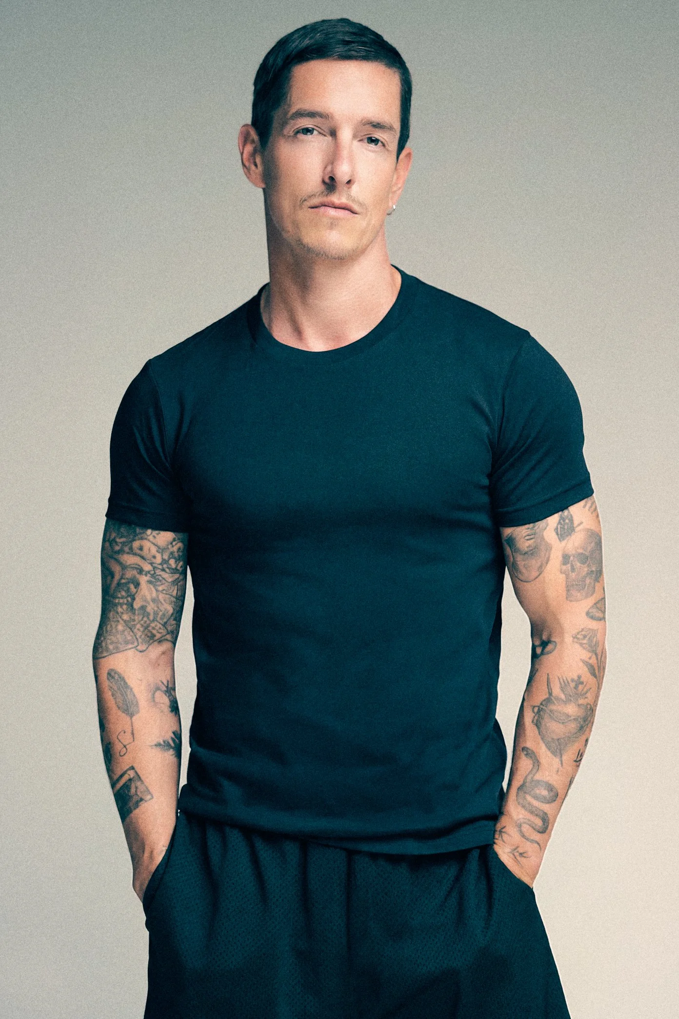 Tattooed man in a fitted black T-shirt and dark trousers standing with his hands in his pockets.